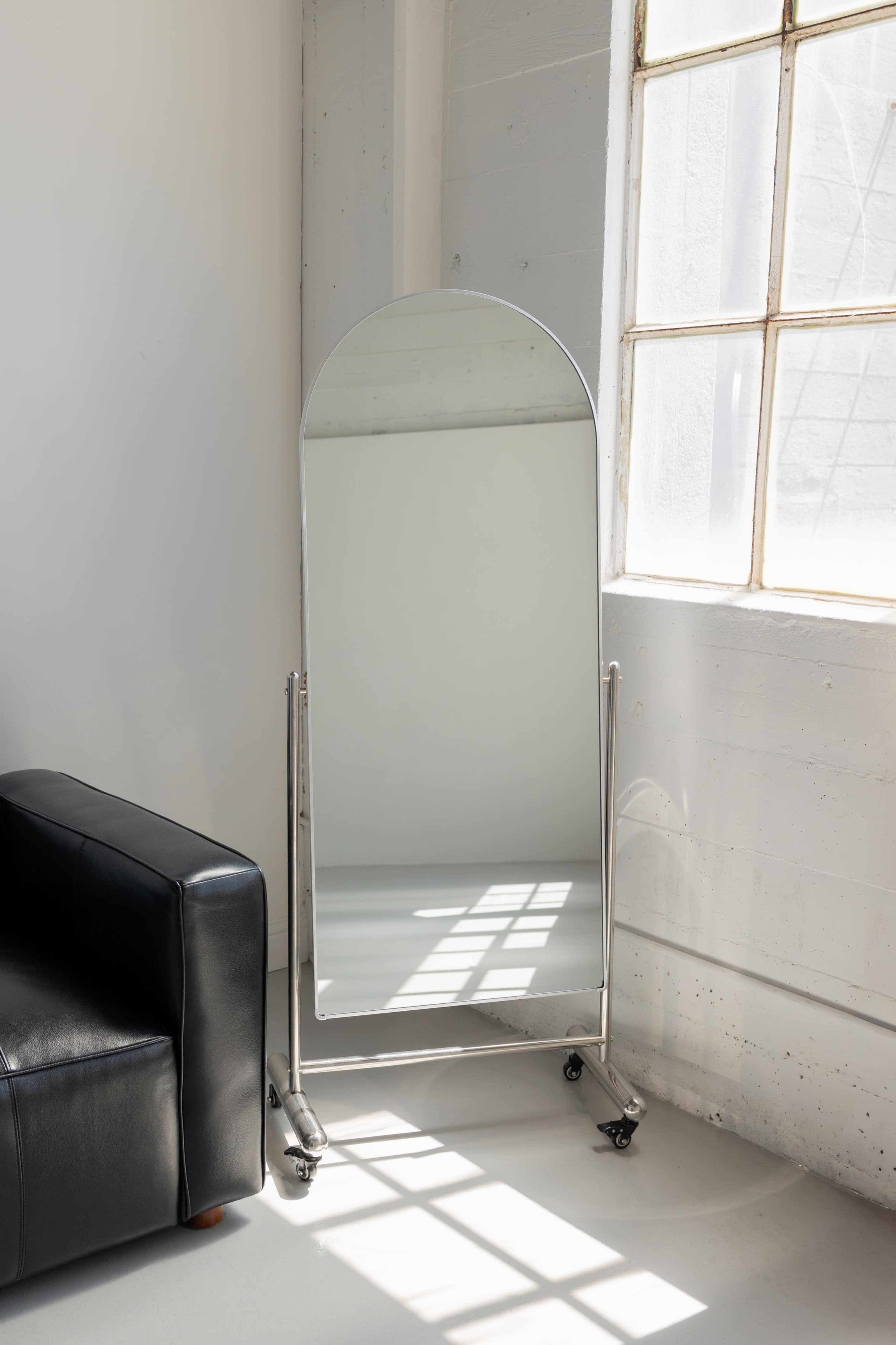 A tall, arched mirror on wheels stands beside a large window with light streaming into a room featuring a black leather sofa.