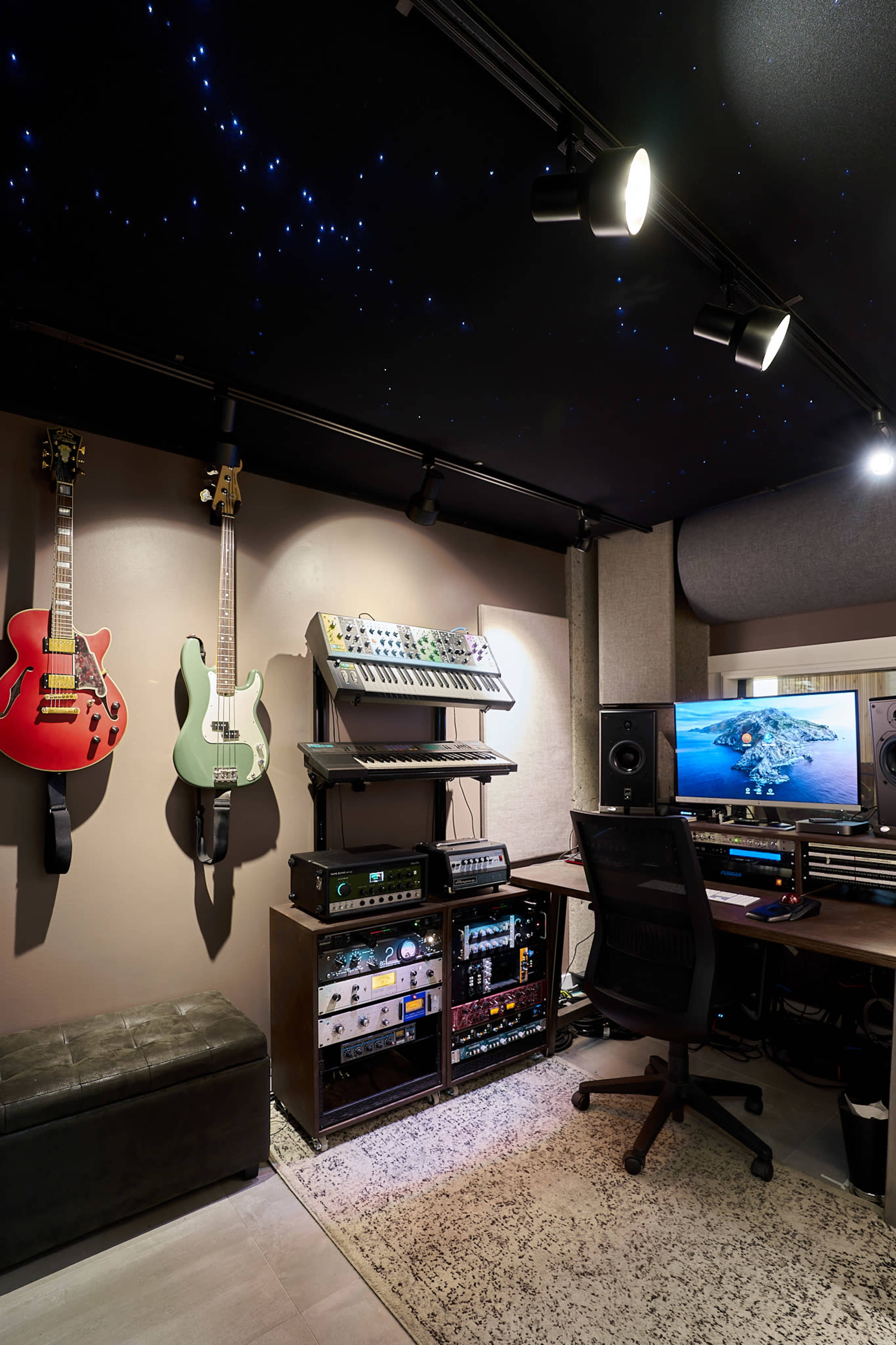 The image shows a compact music production studio with guitars mounted on the wall, audio equipment on a rack, and a computer setup facing a monitor.