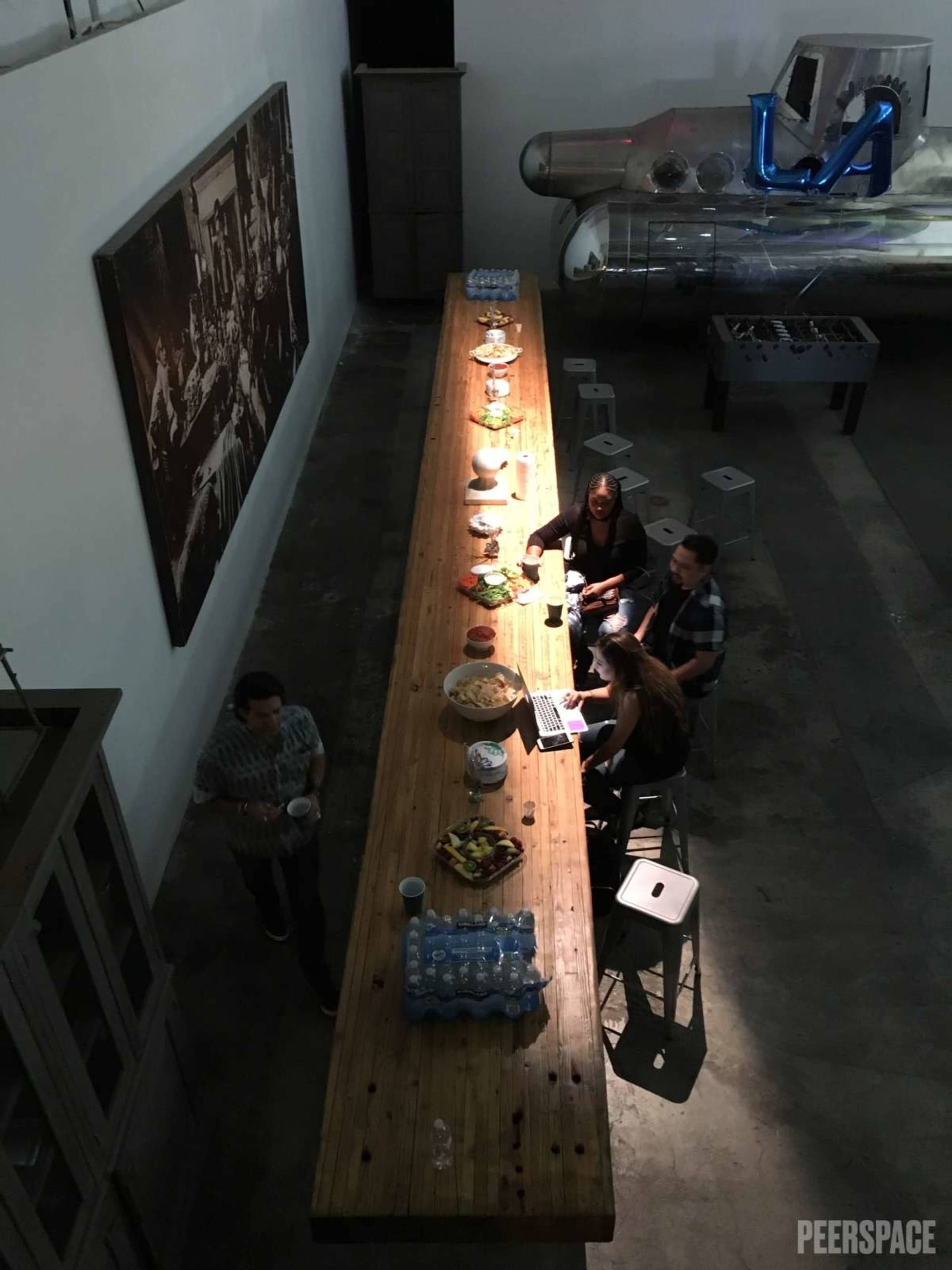 A long wooden table is set with various food items, while four people are seated and engaging with each other and a laptop in a spacious, industrial-style room.