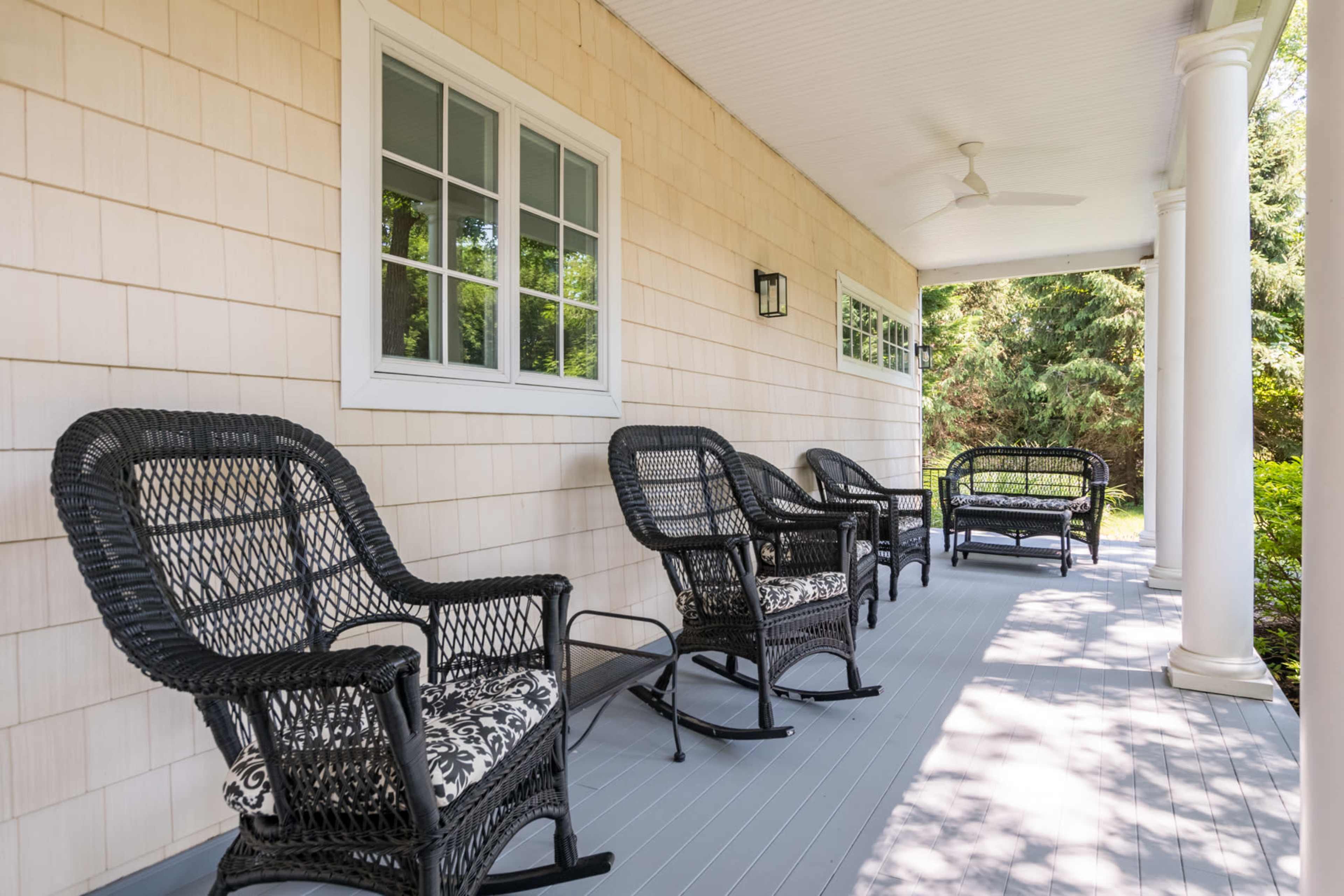 A porch with four black wicker chairs and a ceiling fan, surrounded by greenery.