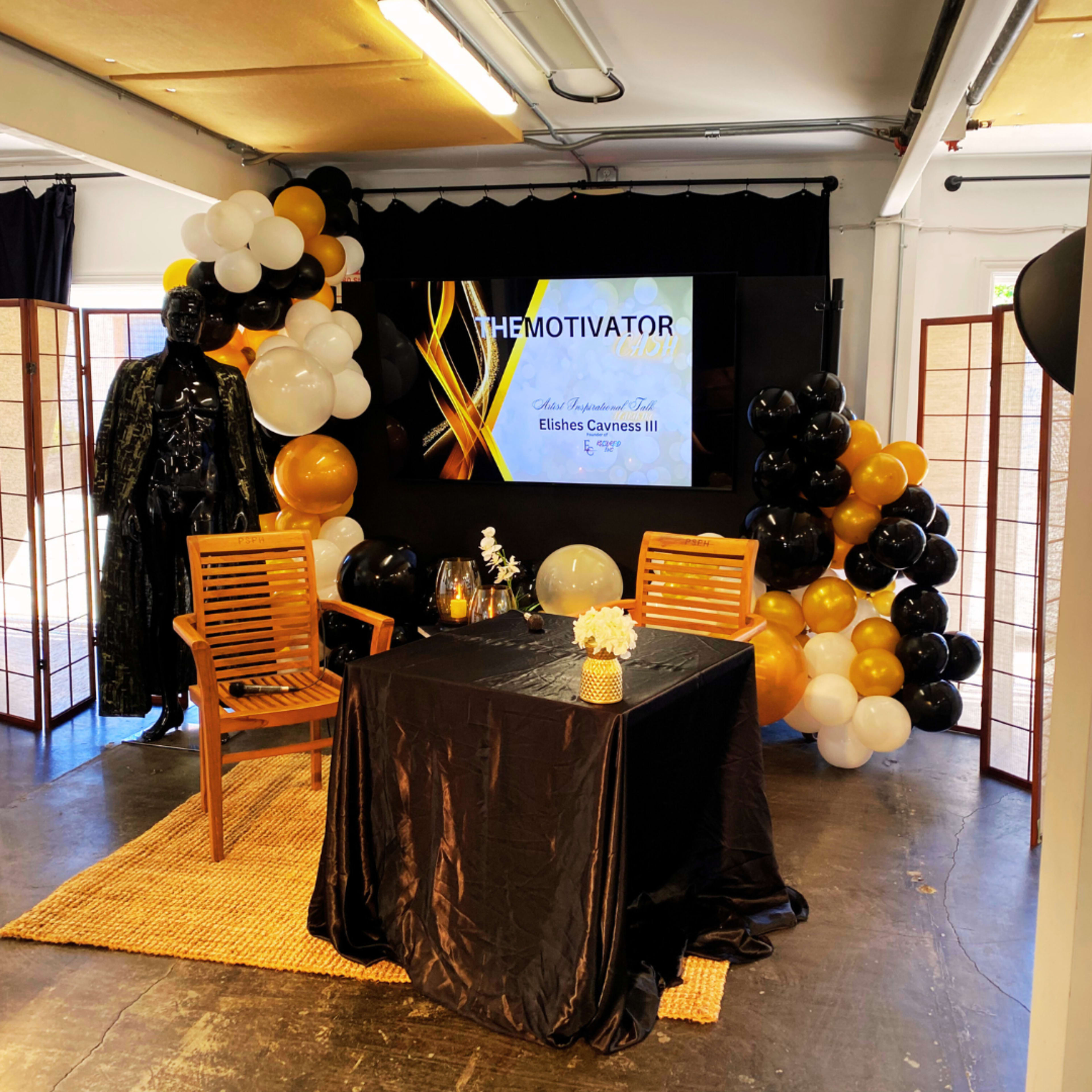 A decorated event space featuring a black and gold balloon arch, a table set with a vase of flowers, two wooden chairs, and a mannequin dressed in a black outfit beside a large screen displaying "THE MOTIVATOR."
