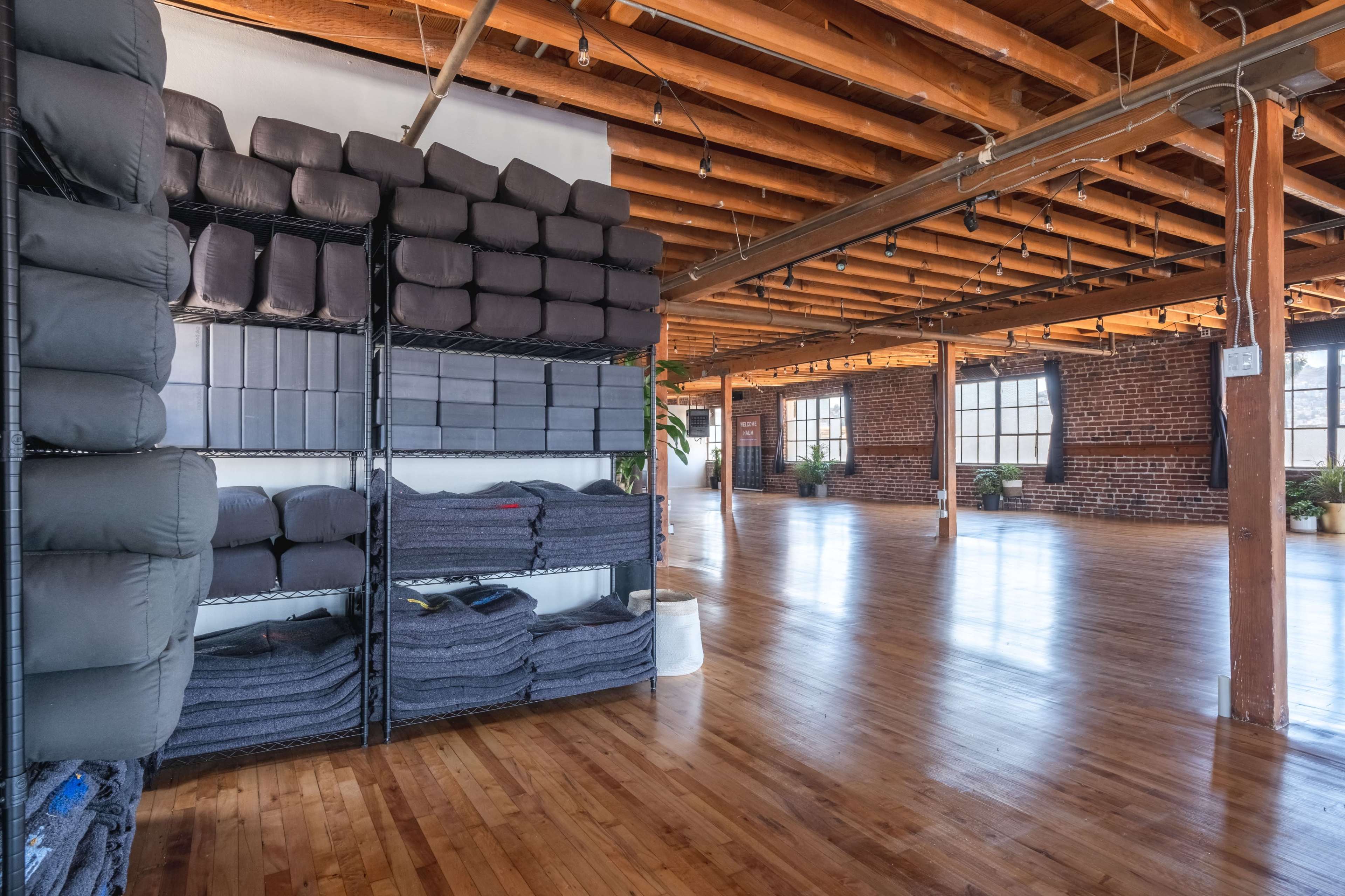 A spacious, minimalist room with wooden floors, large windows, and shelves stacked with cushions and blankets.