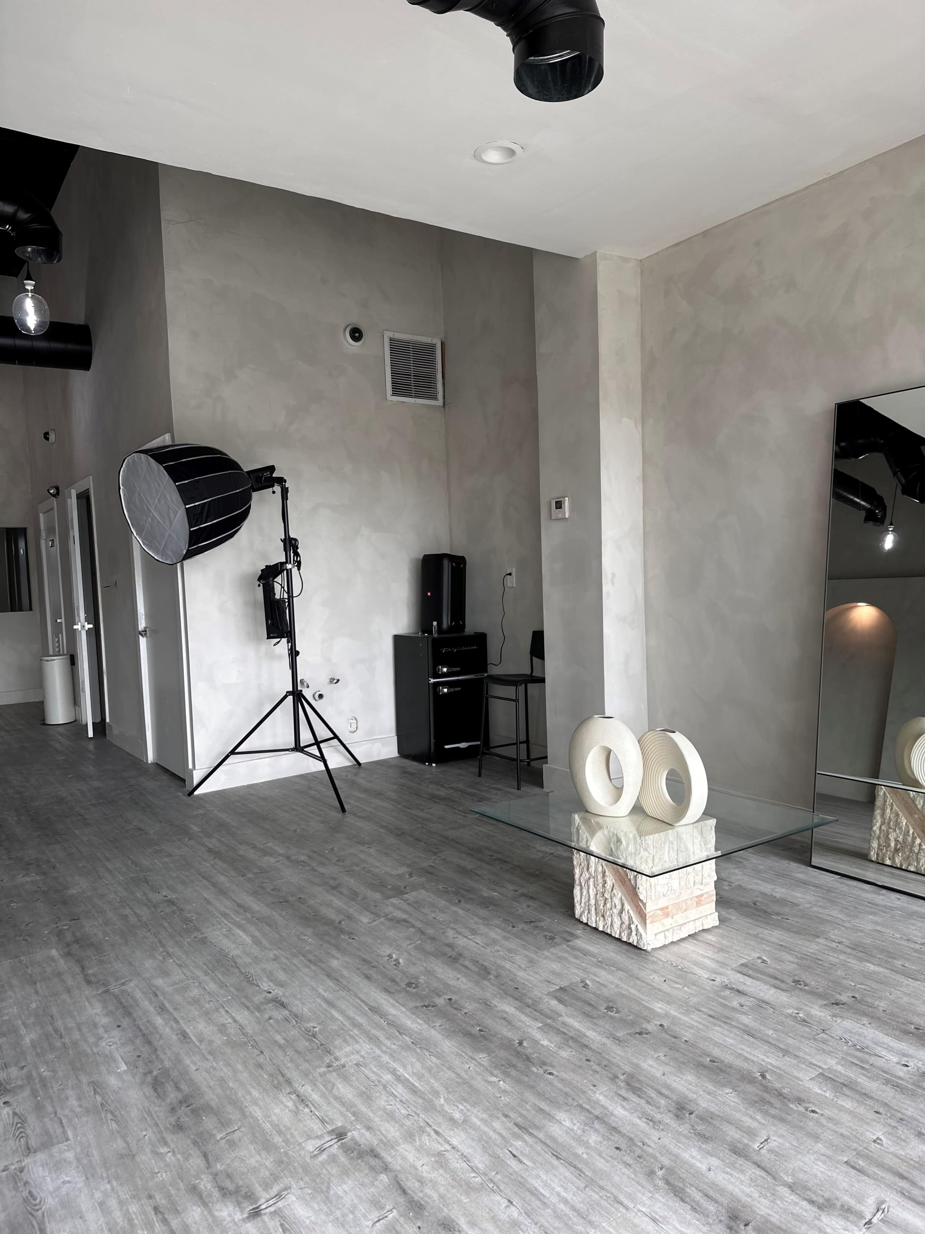 The image shows a minimalist interior space with smooth gray walls, a large mirror, a glass table on a textured pedestal, and studio lighting equipment.