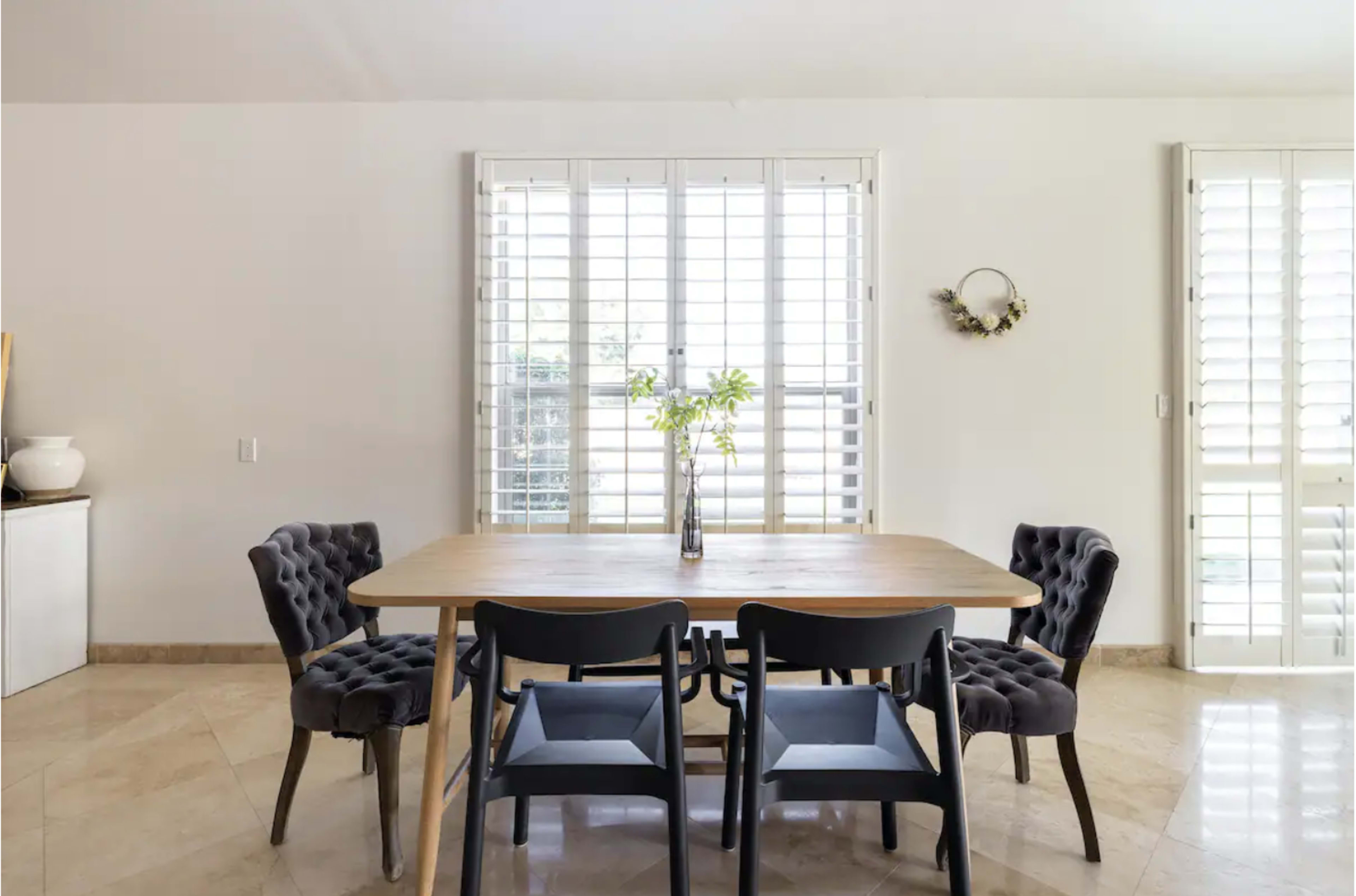 A modern dining area features a wooden table surrounded by upholstered chairs, with light filtering through shuttered windows.