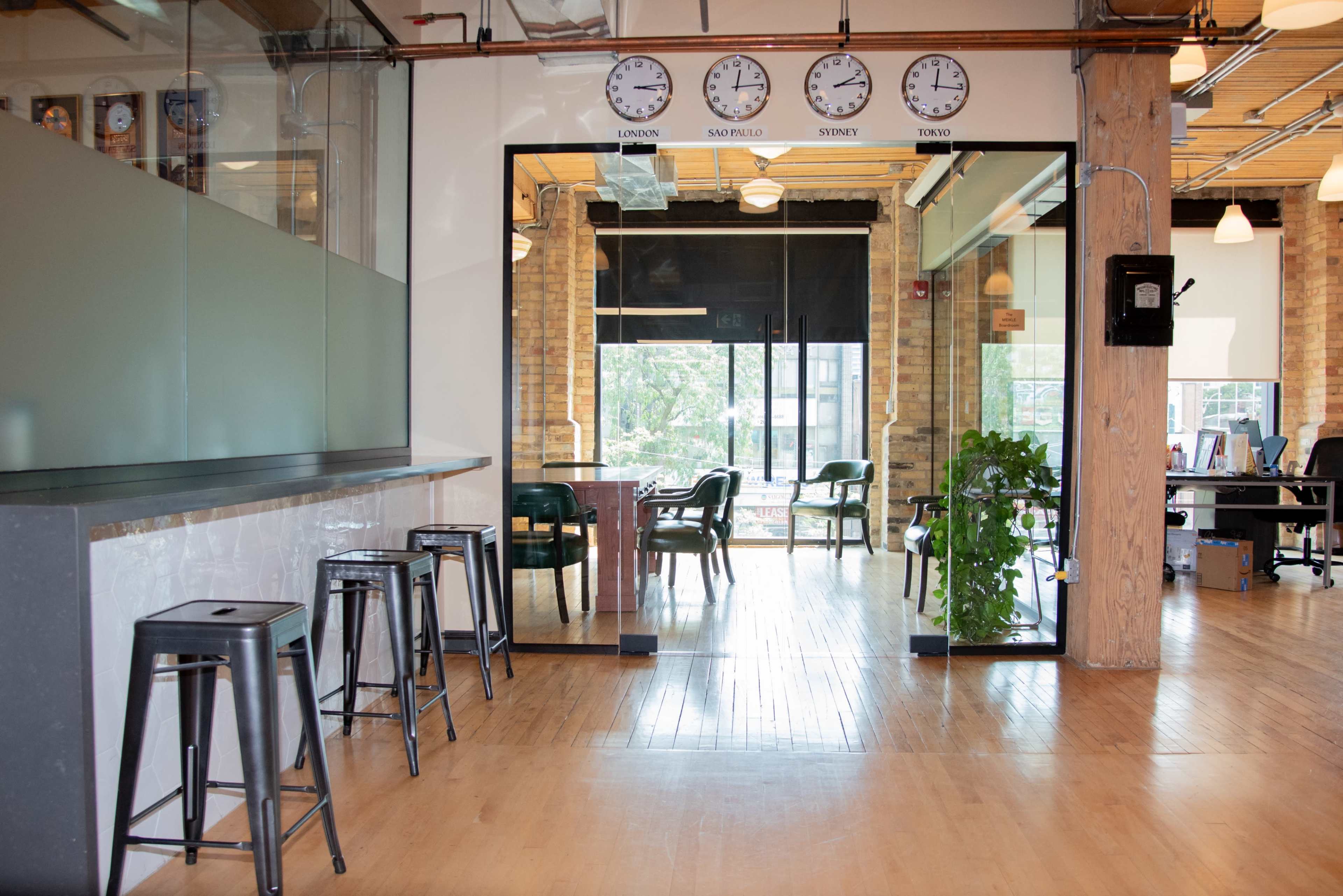 The image shows a modern office space featuring a bar area with stools, a glass partition with clocks displaying different time zones, and a view of a workspace filled with plants and desks.