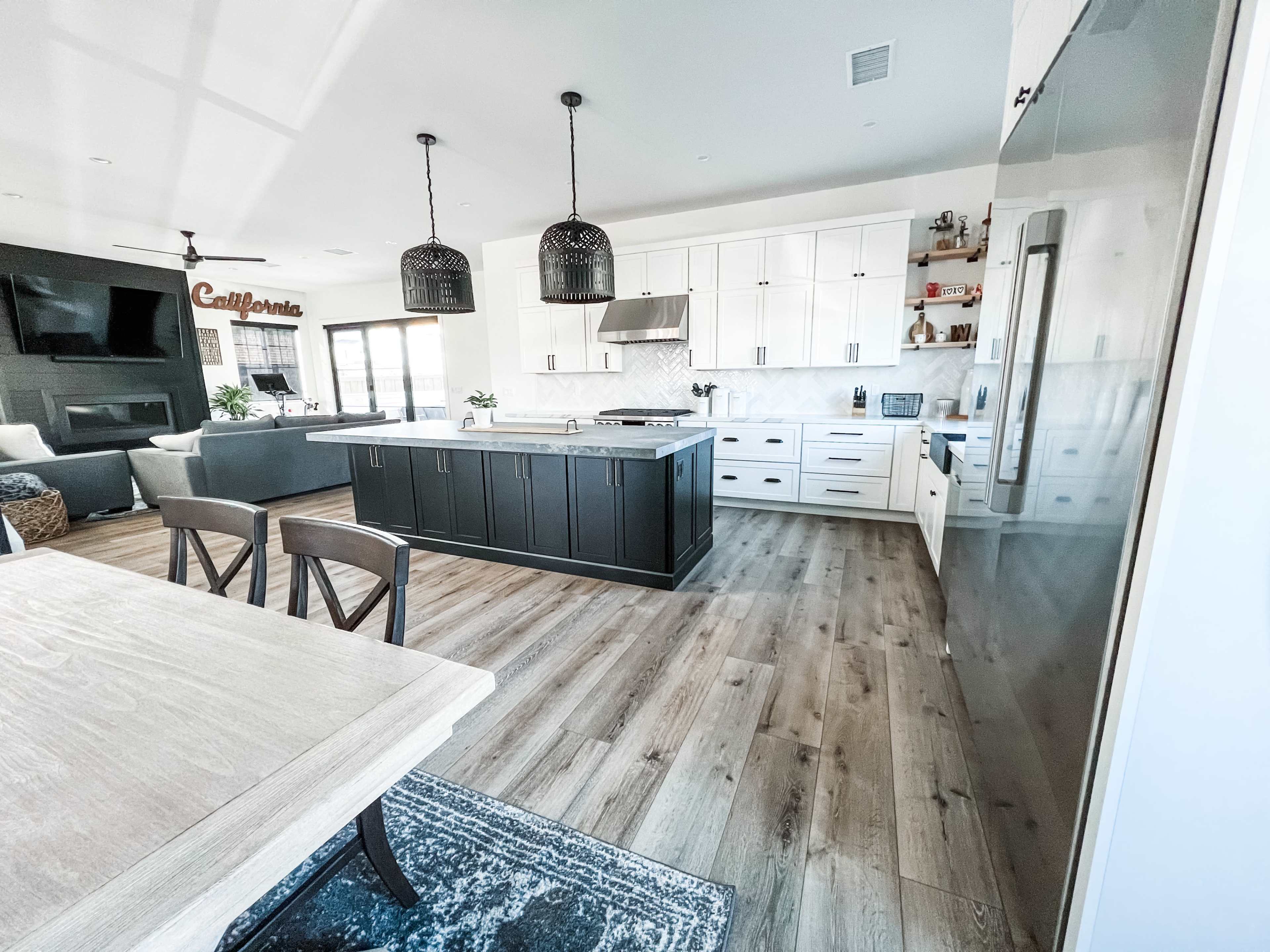 A modern kitchen with white cabinets, a large island, pendant lights, and wooden flooring.