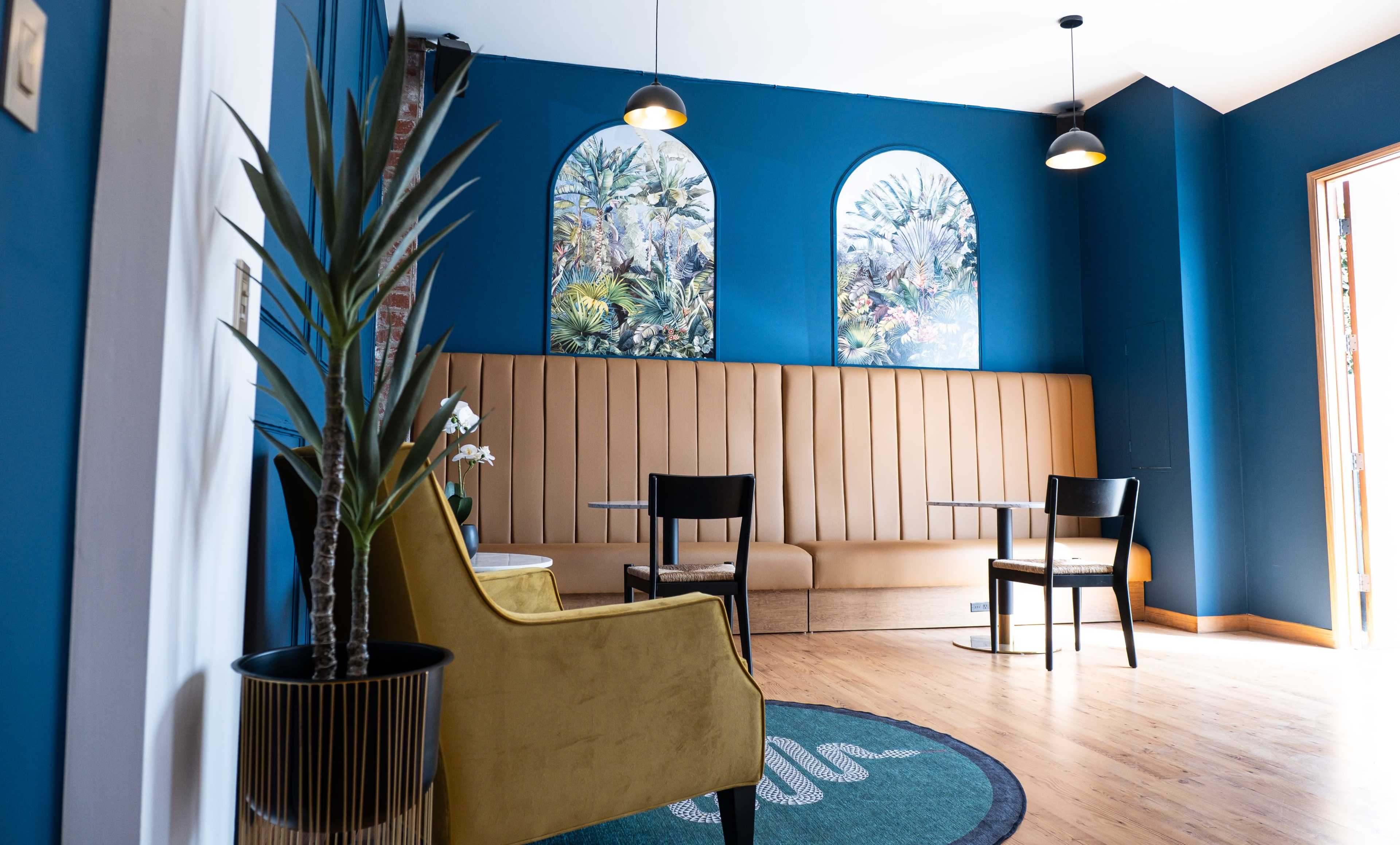 A modern interior with a beige seating area, black chairs, tropical-themed artwork, and a potted plant against deep blue walls.