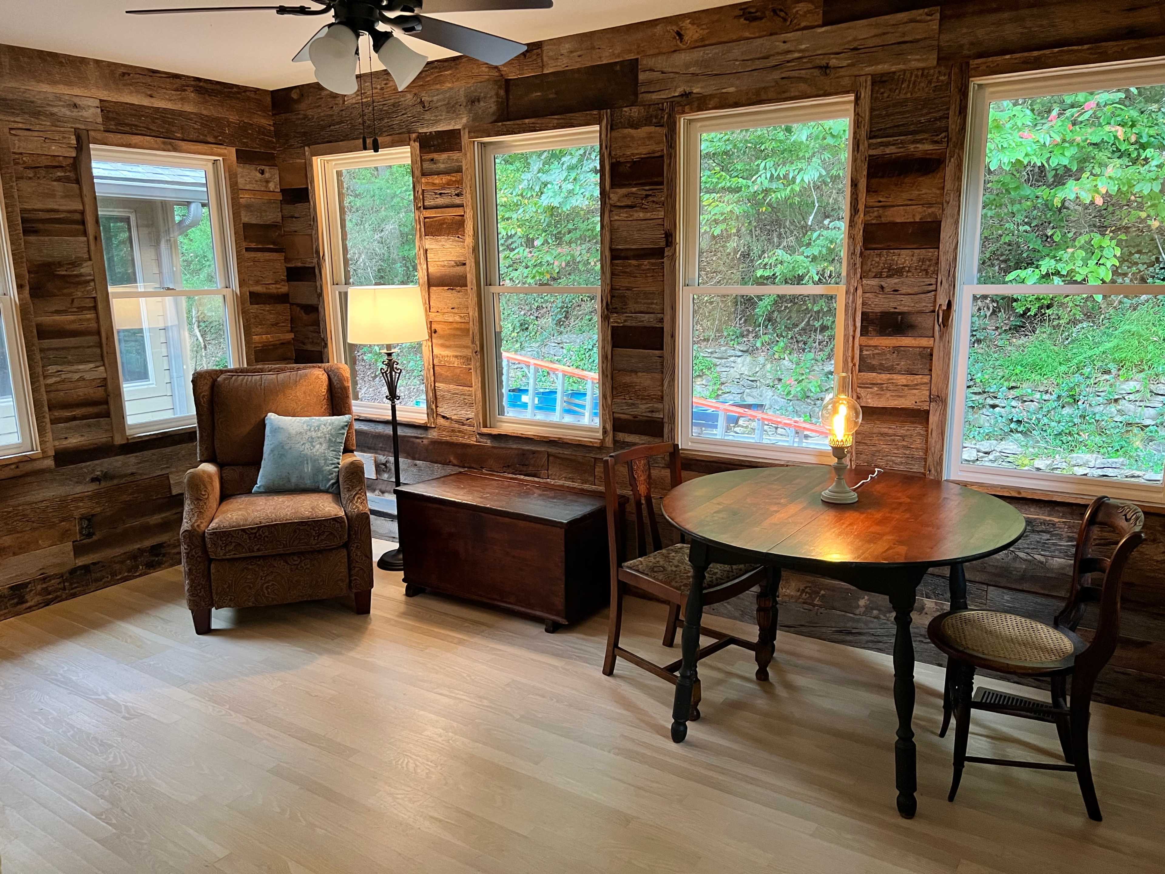 A cozy, rustic room with wooden walls, a table and chairs, an armchair, and a lamp illuminating the space.