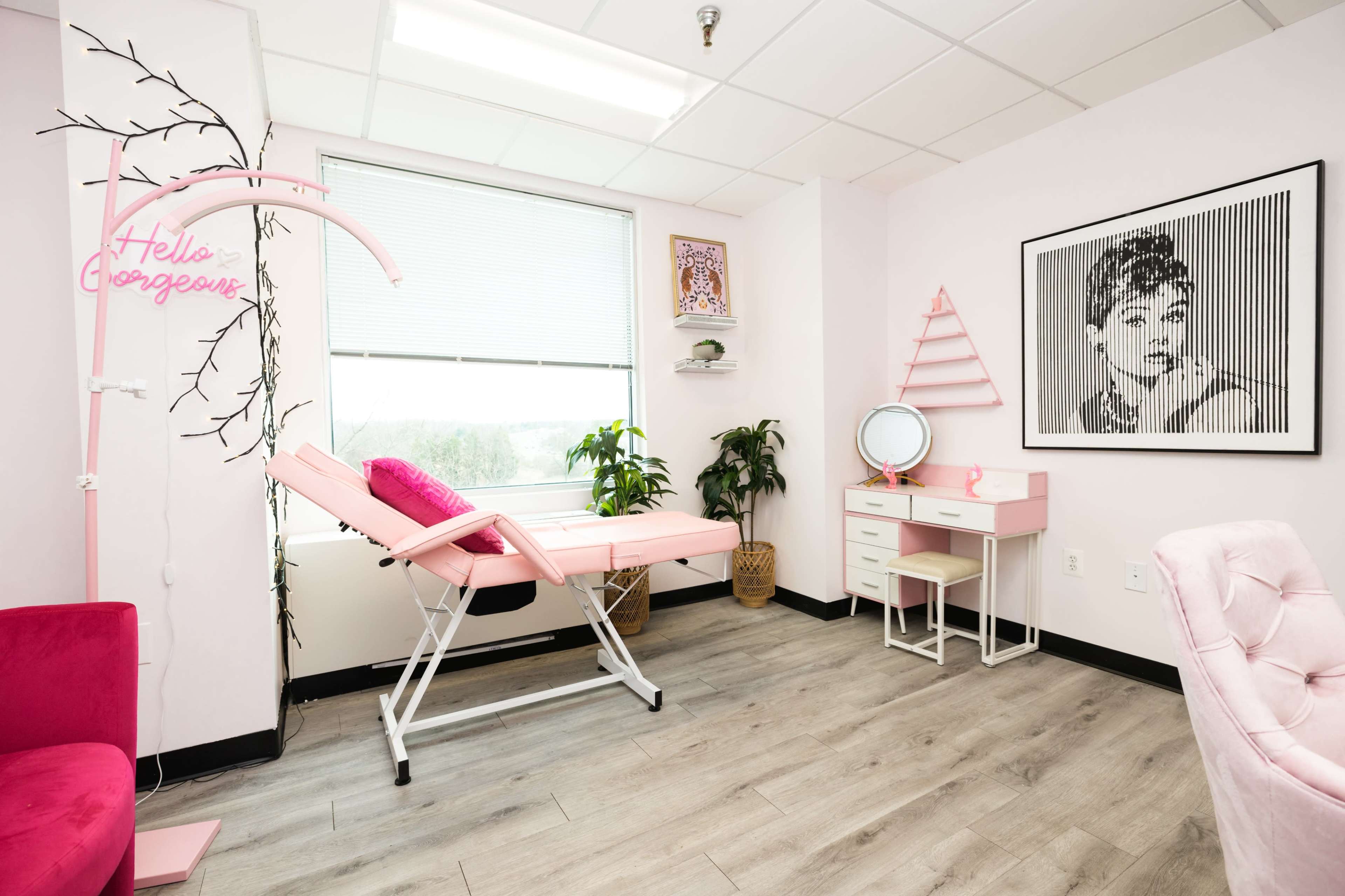 The room features a pink treatment bed, a small vanity desk, and a framed black-and-white portrait on the wall.