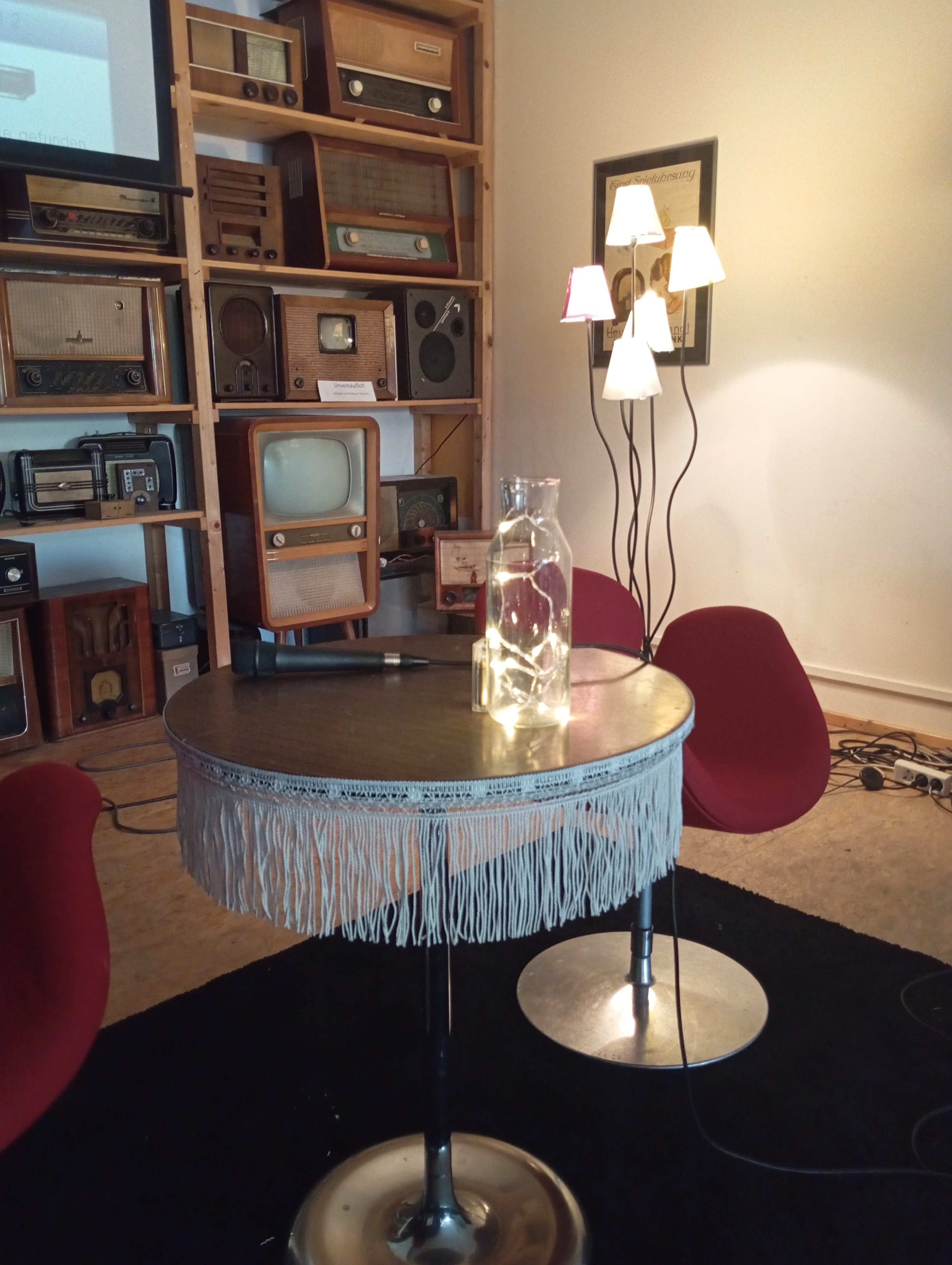 The image shows a cozy setup featuring a round table with a fringed edge, a glass jar filled with lights, two red chairs, and a standing lamp with multiple shades, all against a backdrop of vintage radios.