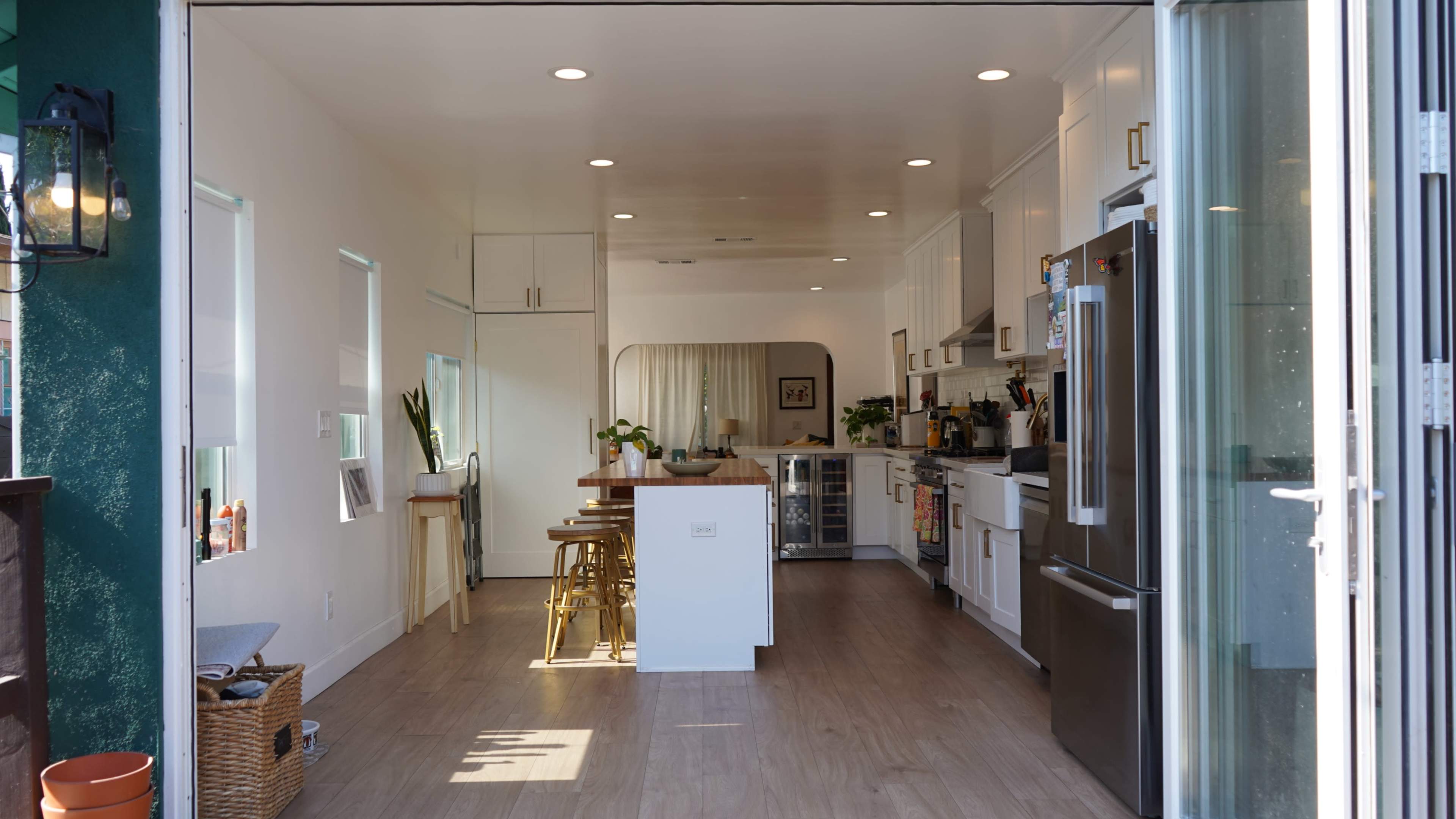 The image shows a modern kitchen featuring white cabinetry, a central island with bar stools, and large windows that open to the outside.