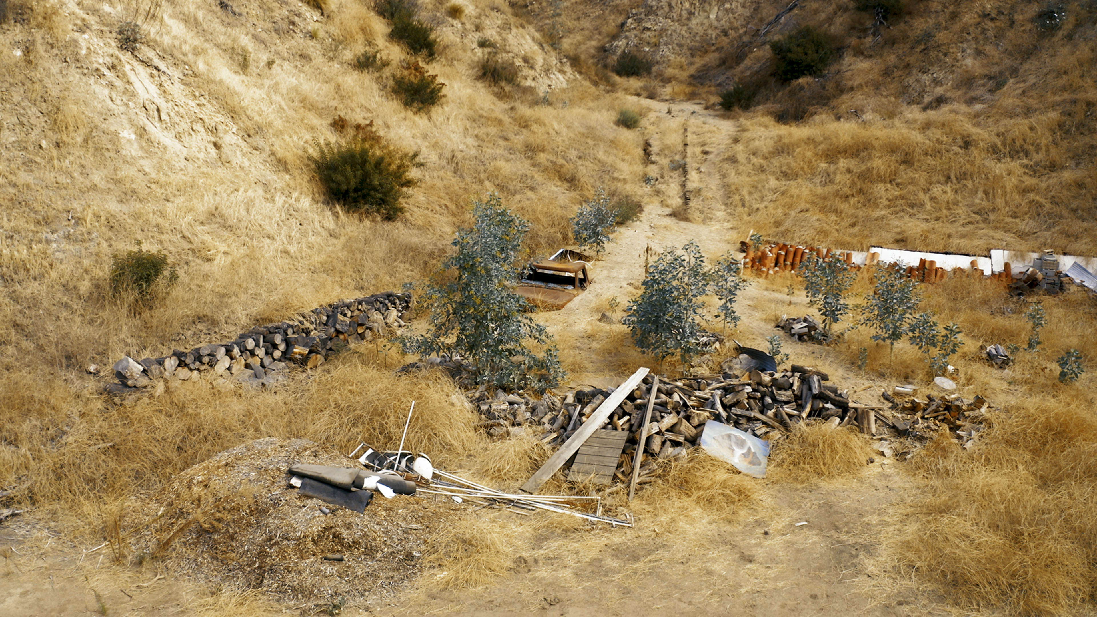 The image shows a dry, uneven landscape with scattered debris, including piles of wood and metal, surrounded by sparse vegetation and rocky ground.