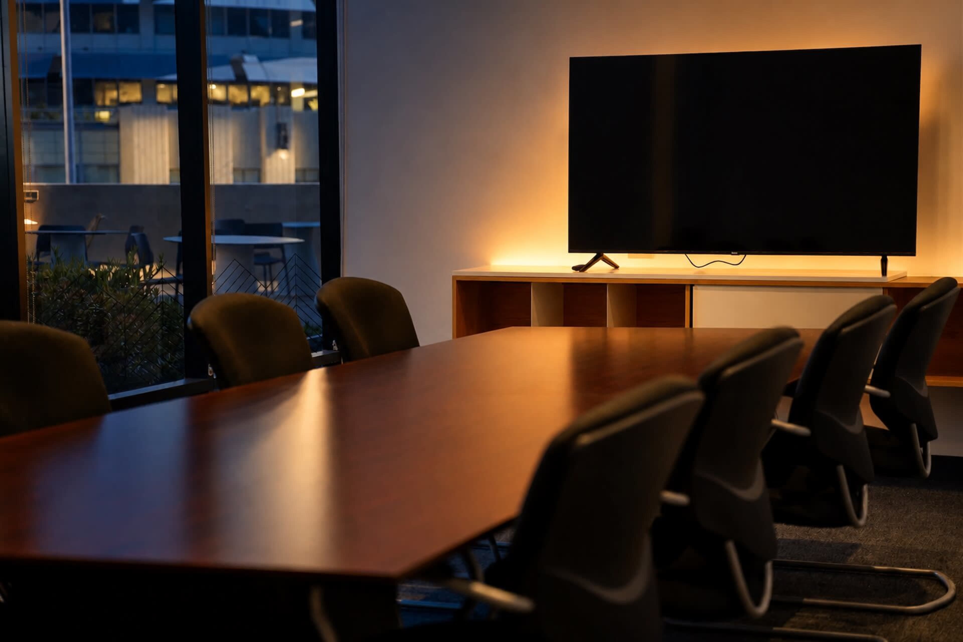 A conference room features a long wooden table surrounded by black chairs, with a television mounted on a wall illuminated by soft lighting.