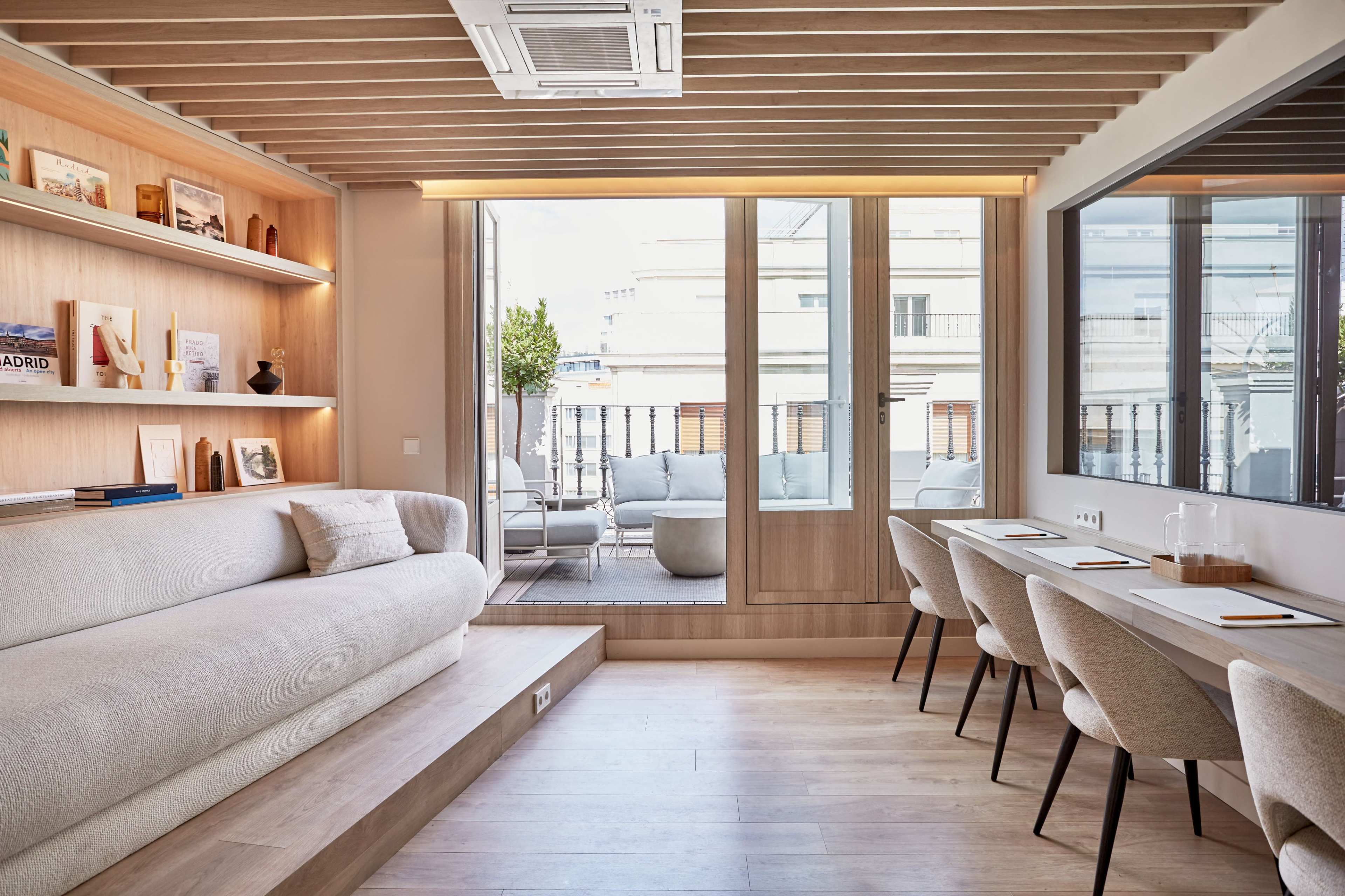 The image shows a modern interior living space featuring a light-colored sofa, wooden accents, and large windows that open to a balcony.