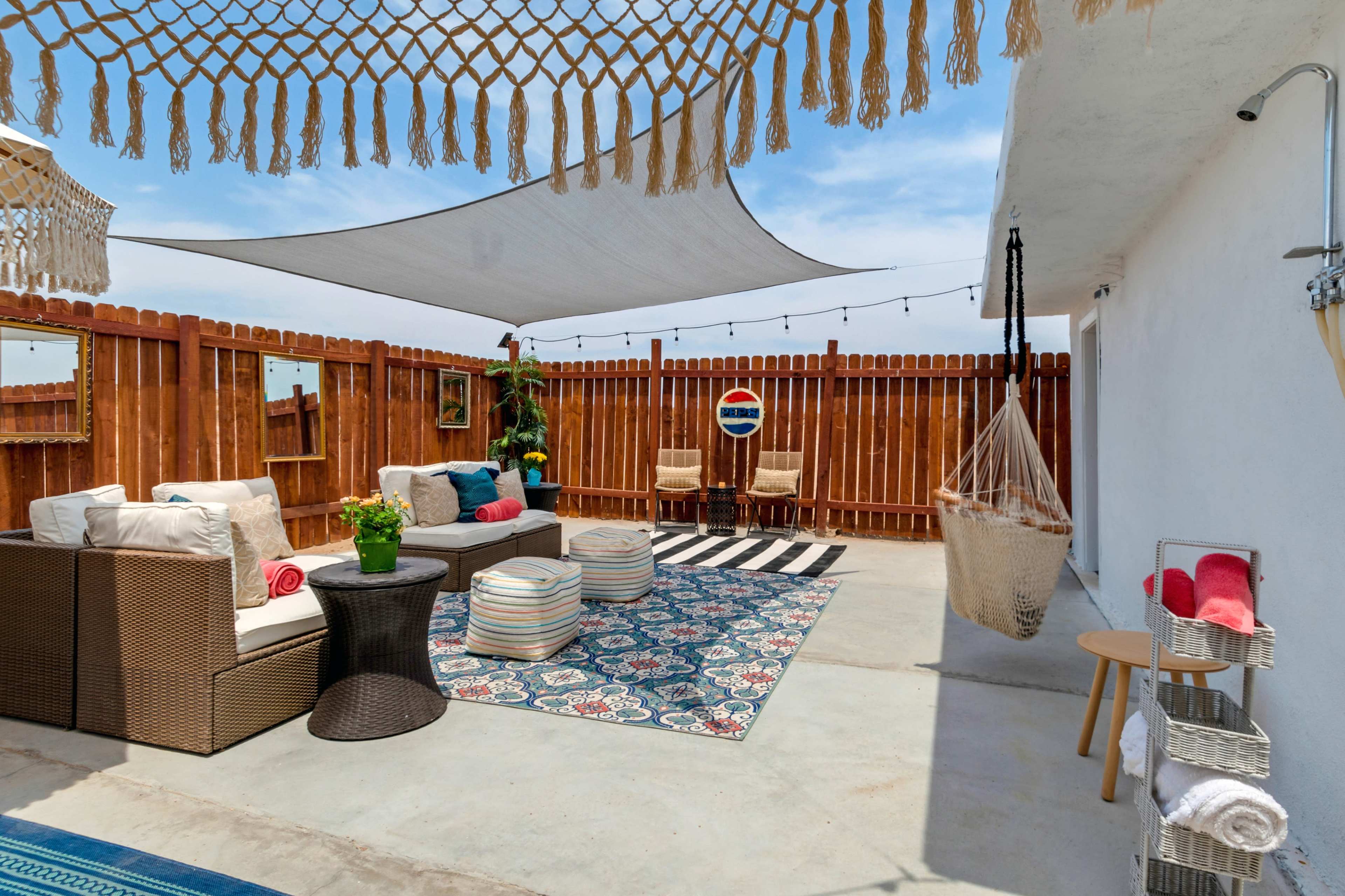 The image depicts a patio area featuring outdoor furniture, a large rug, and decorative elements, all surrounded by wooden fencing and a shade canopy.