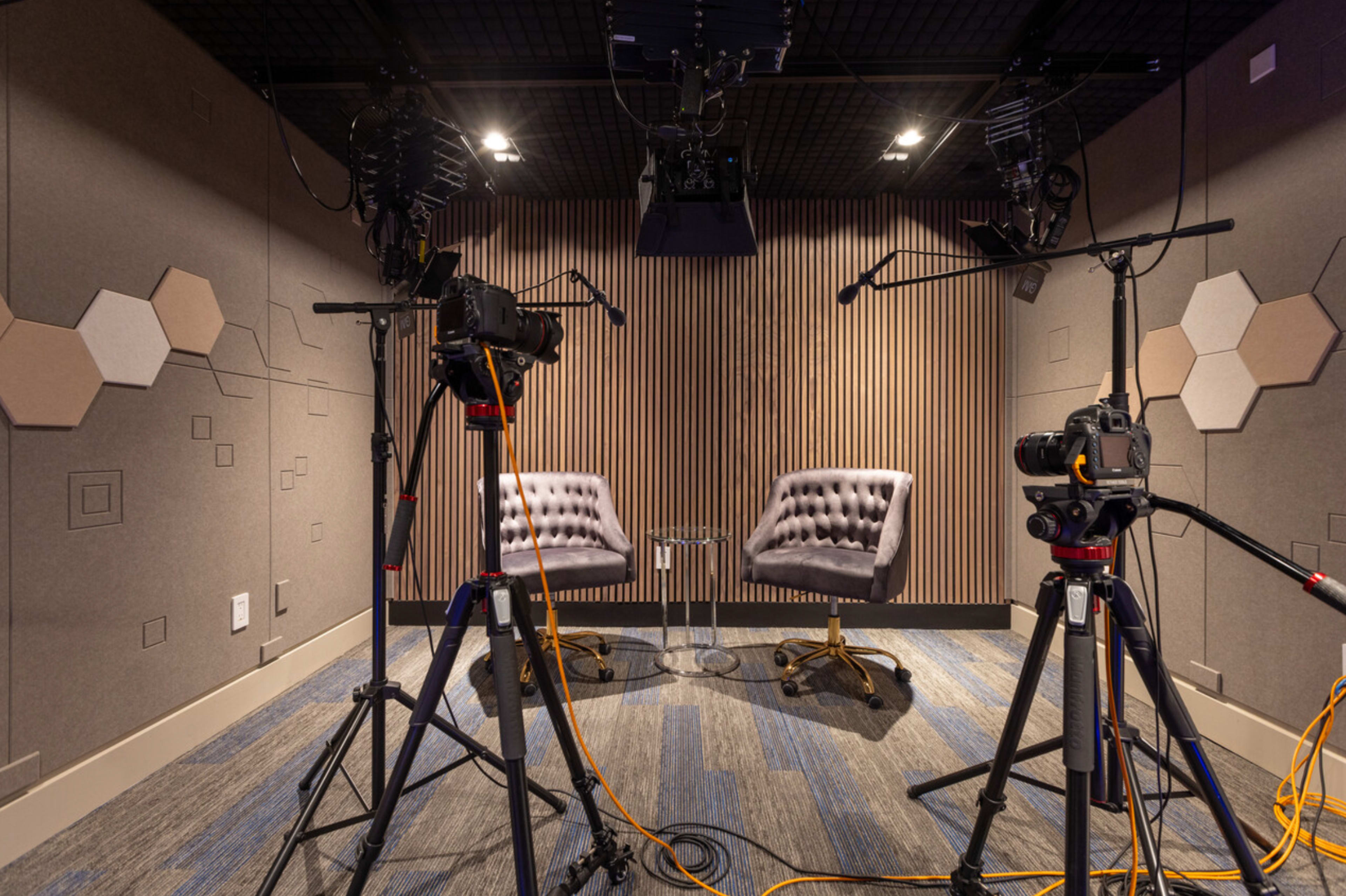 A video recording studio features two tufted chairs positioned in front of a wall with acoustic panels, surrounded by camera equipment and lighting.
