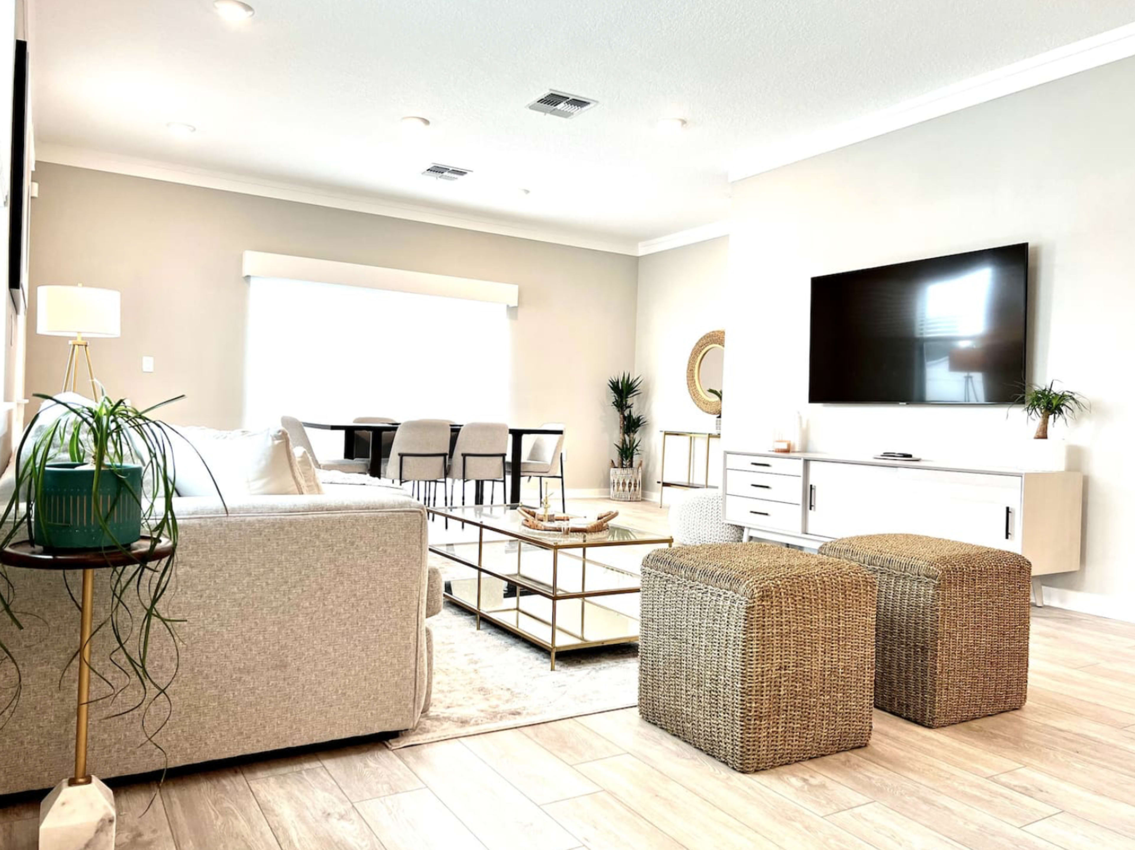 A modern living room features a light-colored sofa, a coffee table, two woven ottomans, and a large TV mounted on the wall.