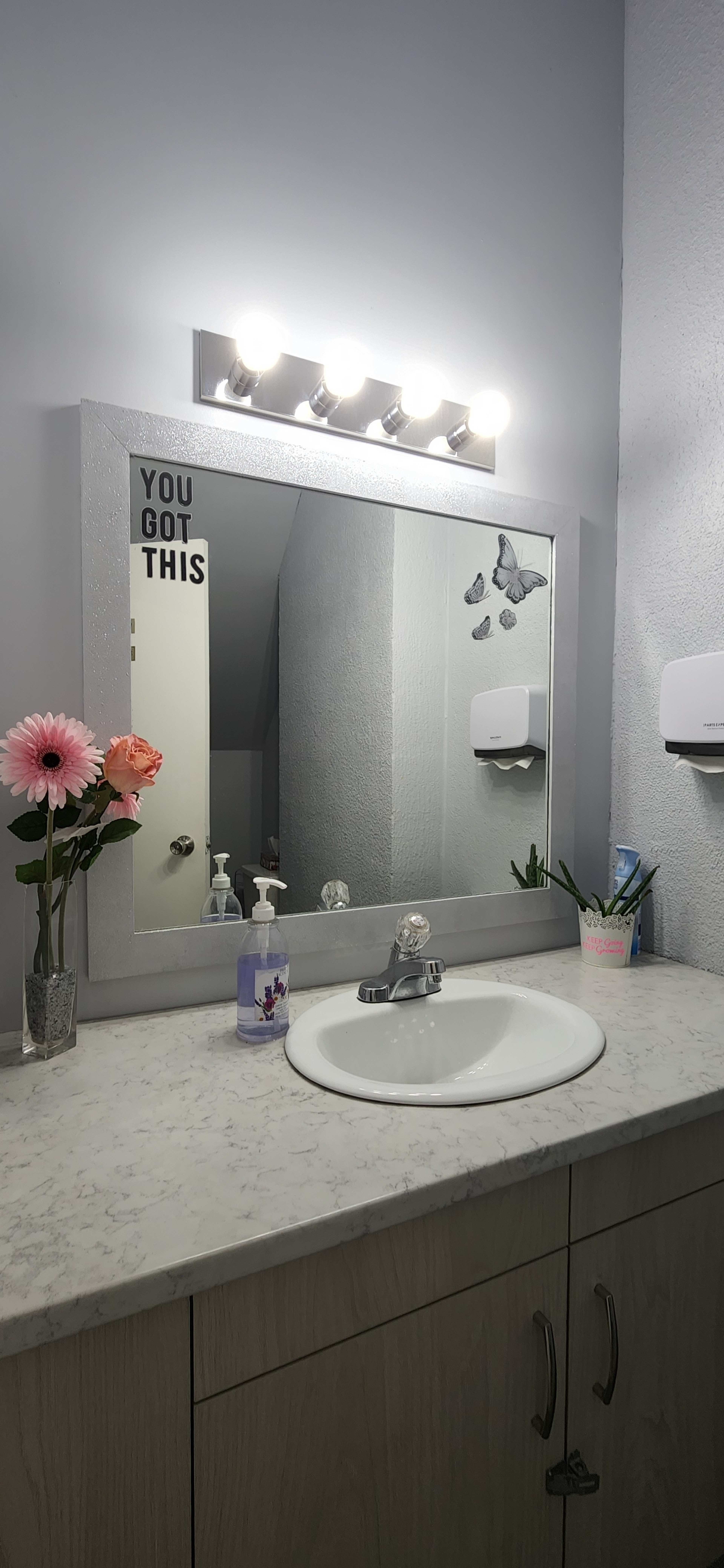 The image shows a bathroom countertop with a sink, a mirror above it, a flower vase, and soap dispensers on the wall.