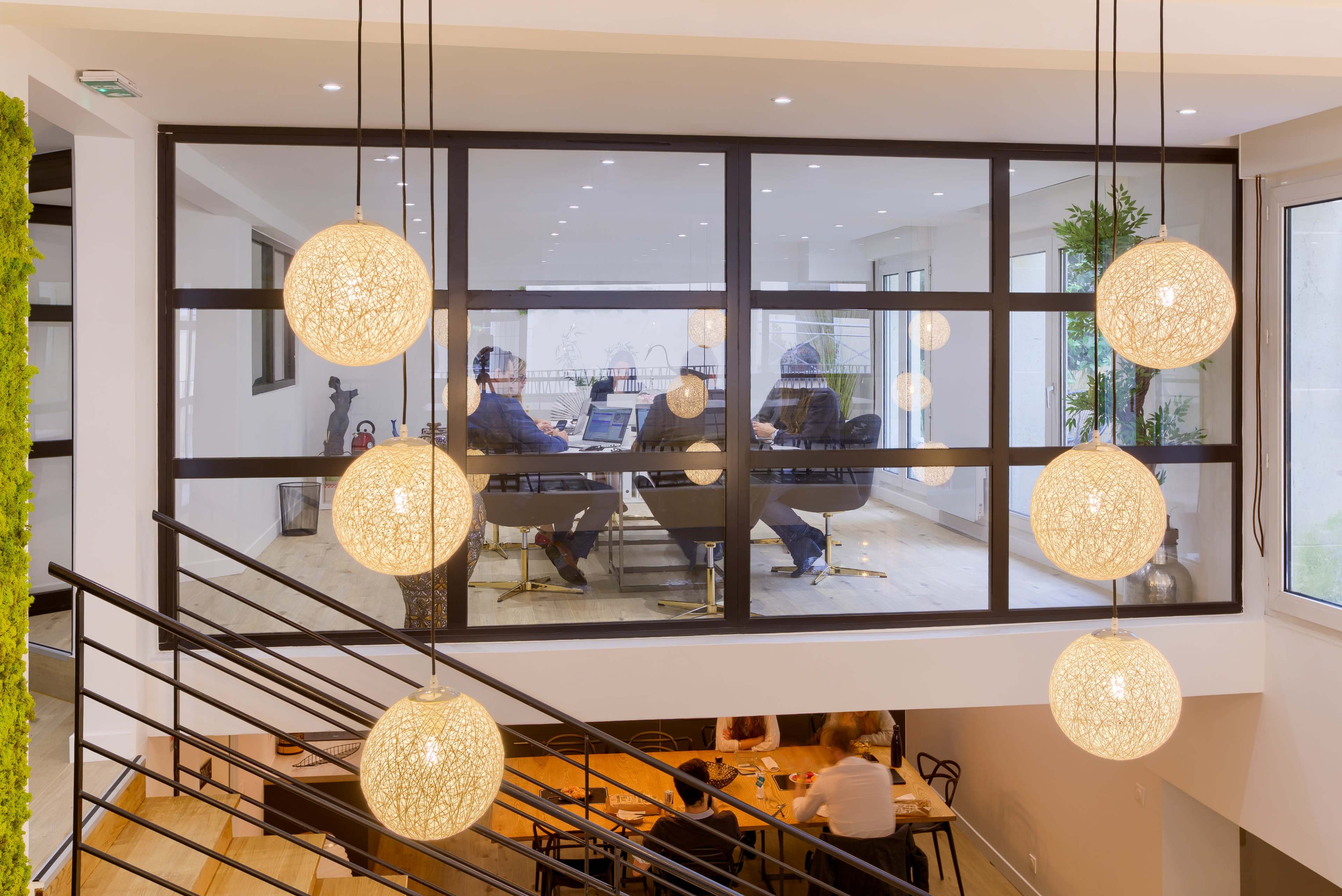 A modern office space features a glass-enclosed meeting room where individuals are seated around a table, illuminated by decorative hanging lights above.