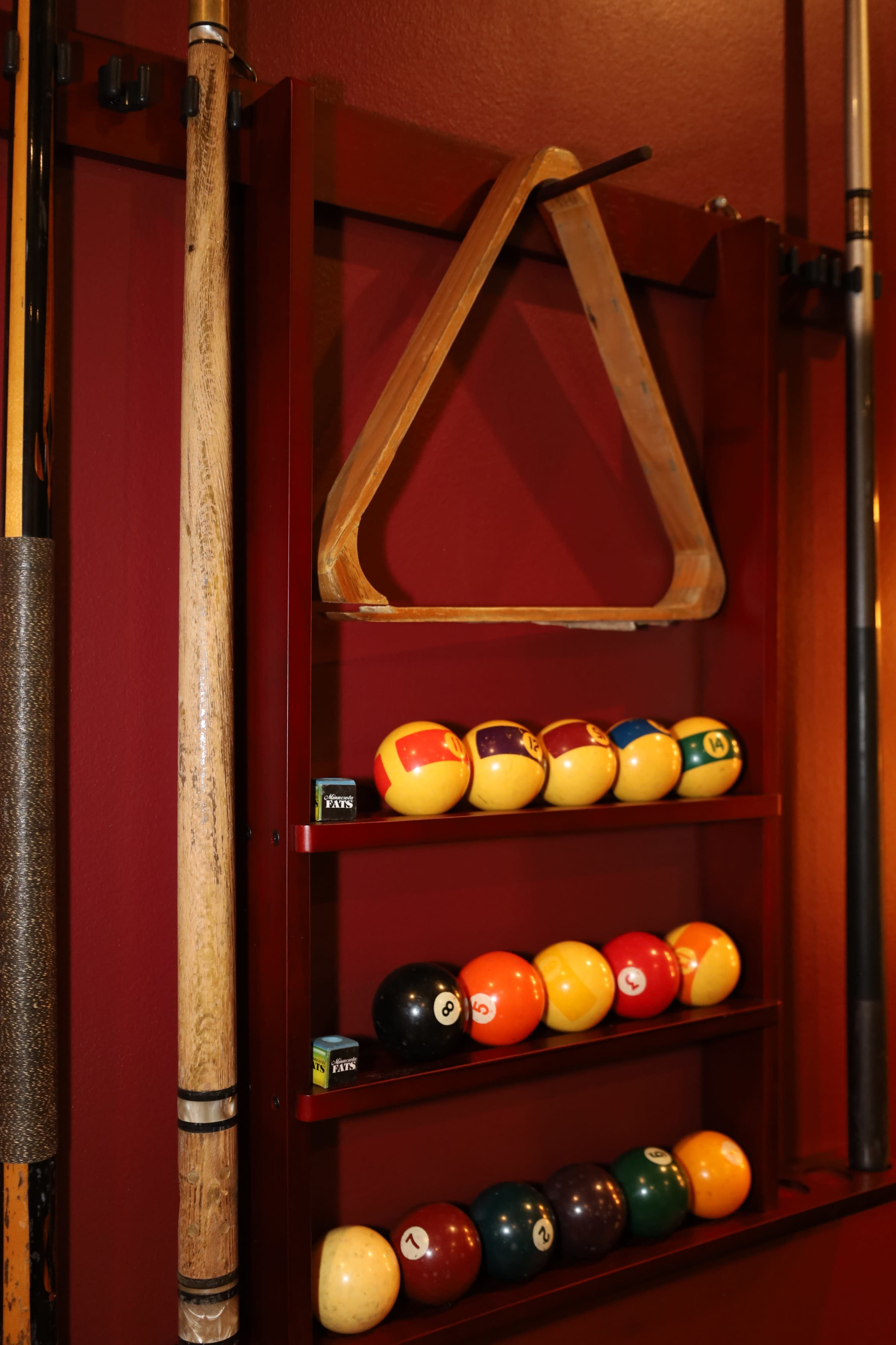 The image shows a rack with a triangular pool table triangle and two rows of billiard balls organized on a red wall.