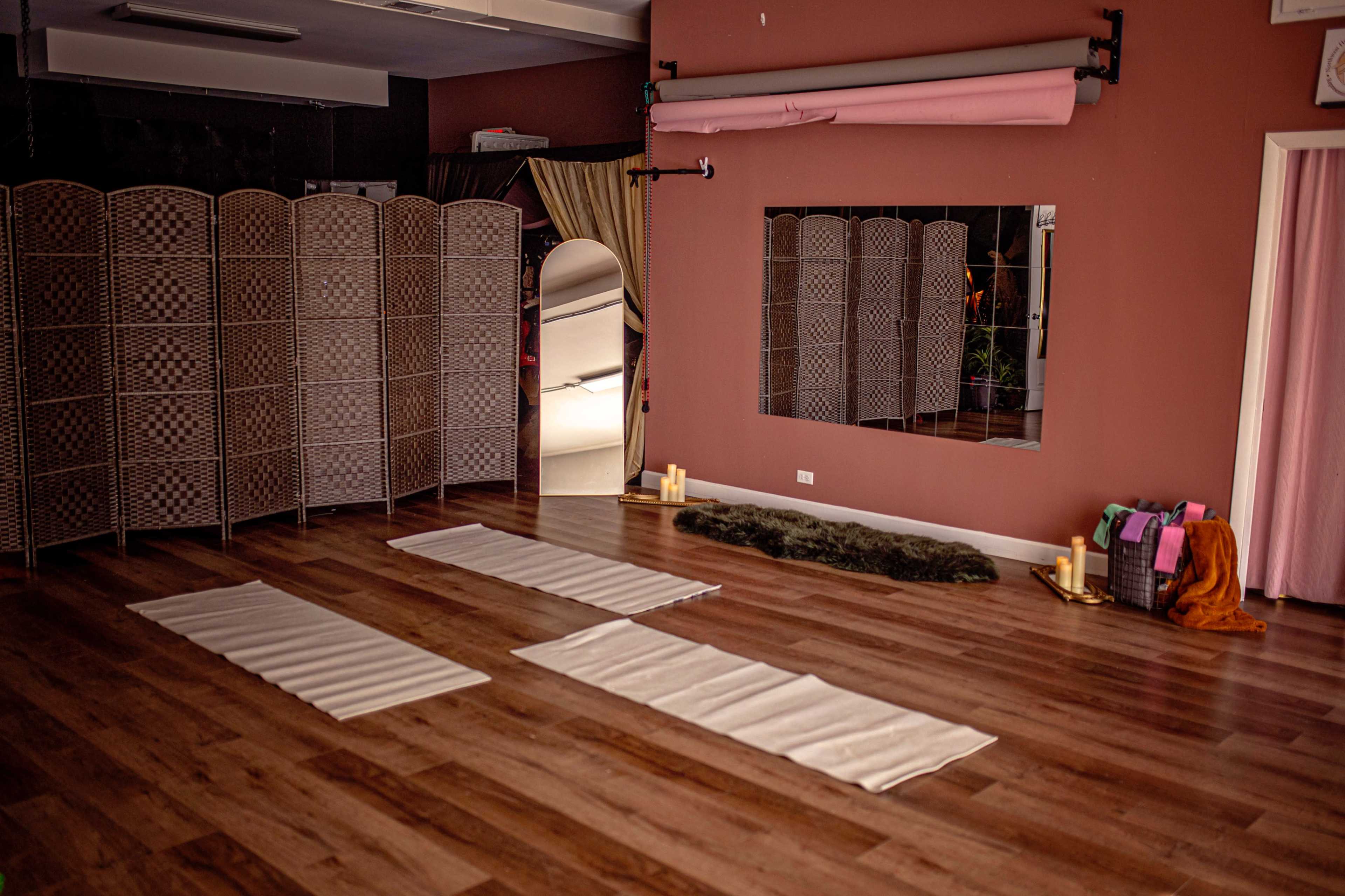 The image shows a spacious room with wooden flooring, three white mats laid out on the floor, a mirrored wall, and a folding room divider.