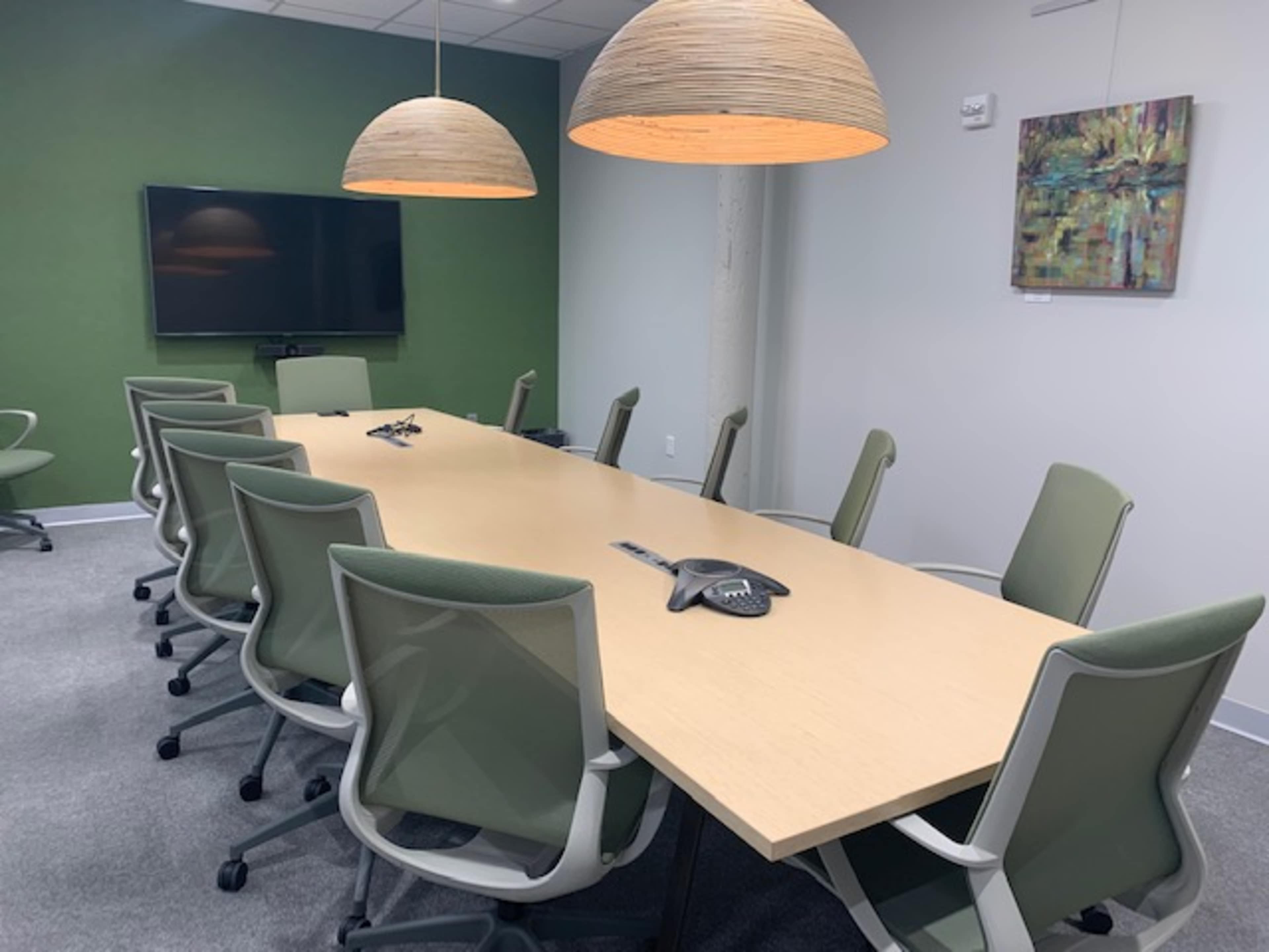 A long wooden conference table with eight green office chairs is positioned under two pendant lights in a modern meeting room adorned with a green accent wall.