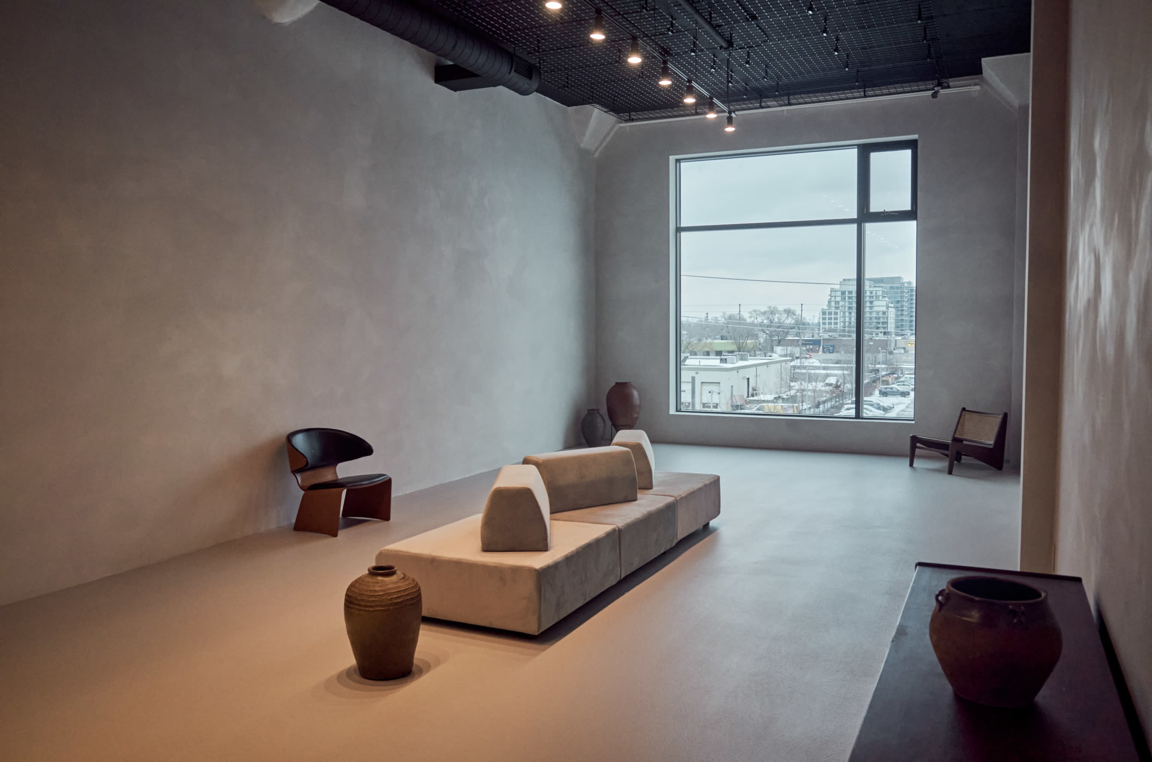 The image shows a minimalist interior space with large windows, featuring a sofa in the center, a chair to the left, and several ceramic pots on the floor.