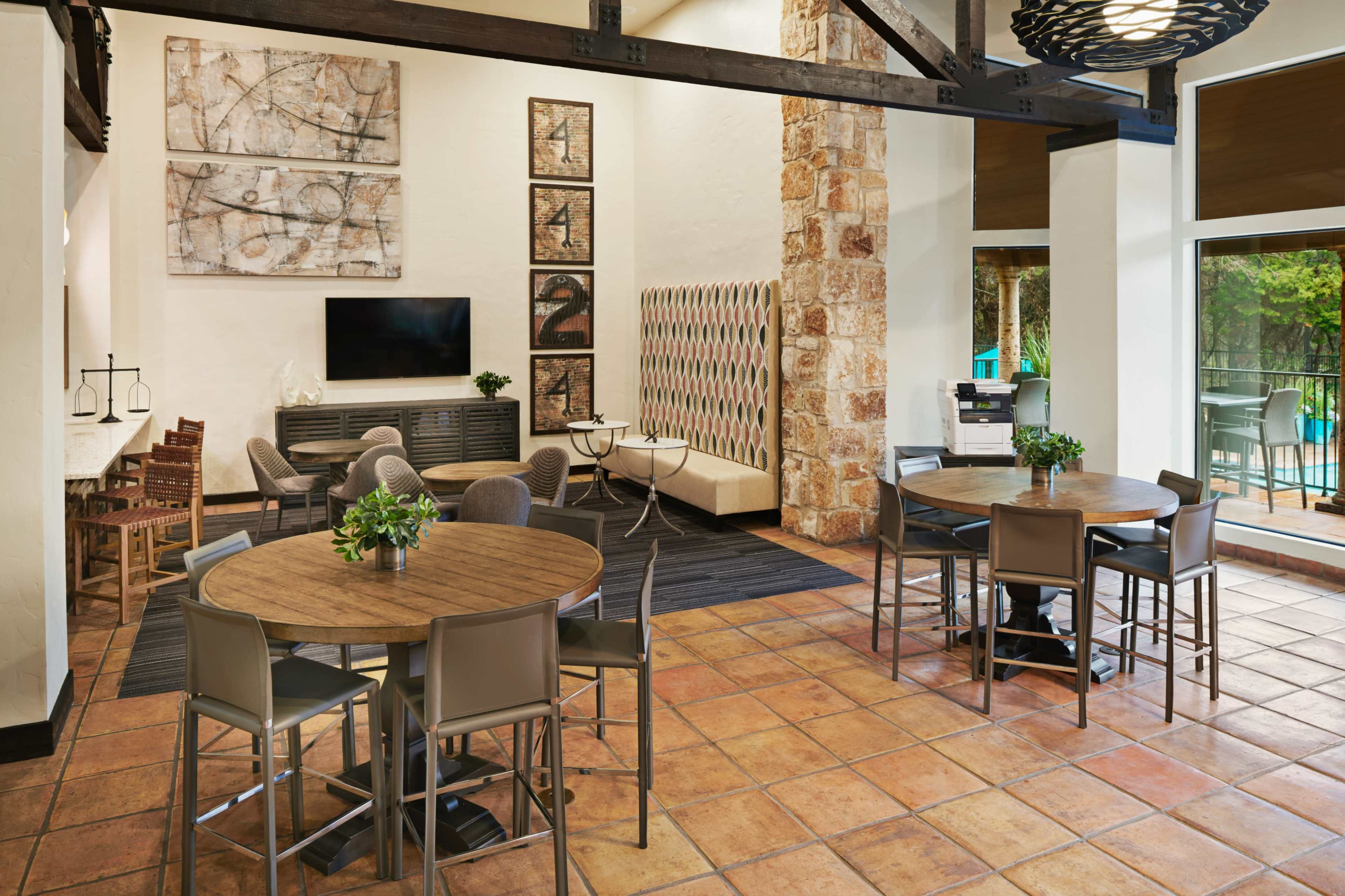 A spacious communal area featuring multiple seating arrangements, including round tables and chairs, with a large wall-mounted TV and decorative artwork.