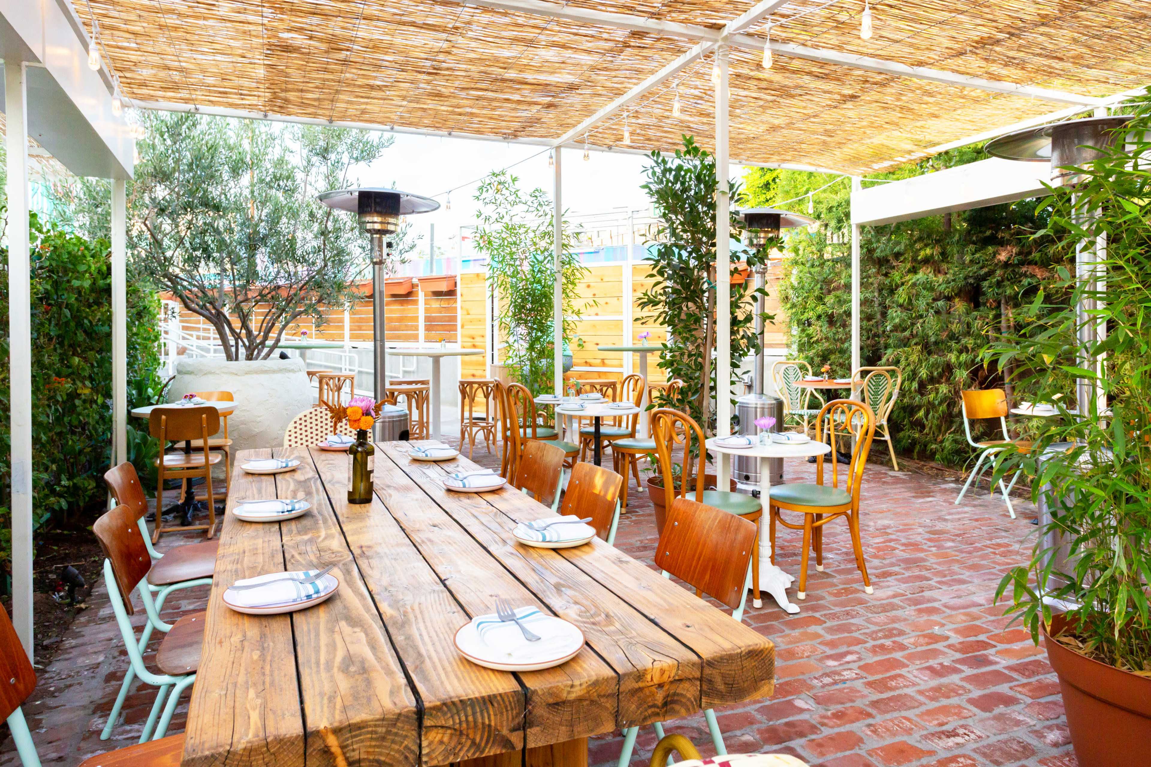 The image shows an outdoor dining area with wooden tables and chairs, surrounded by greenery and patio heaters under a straw canopy.