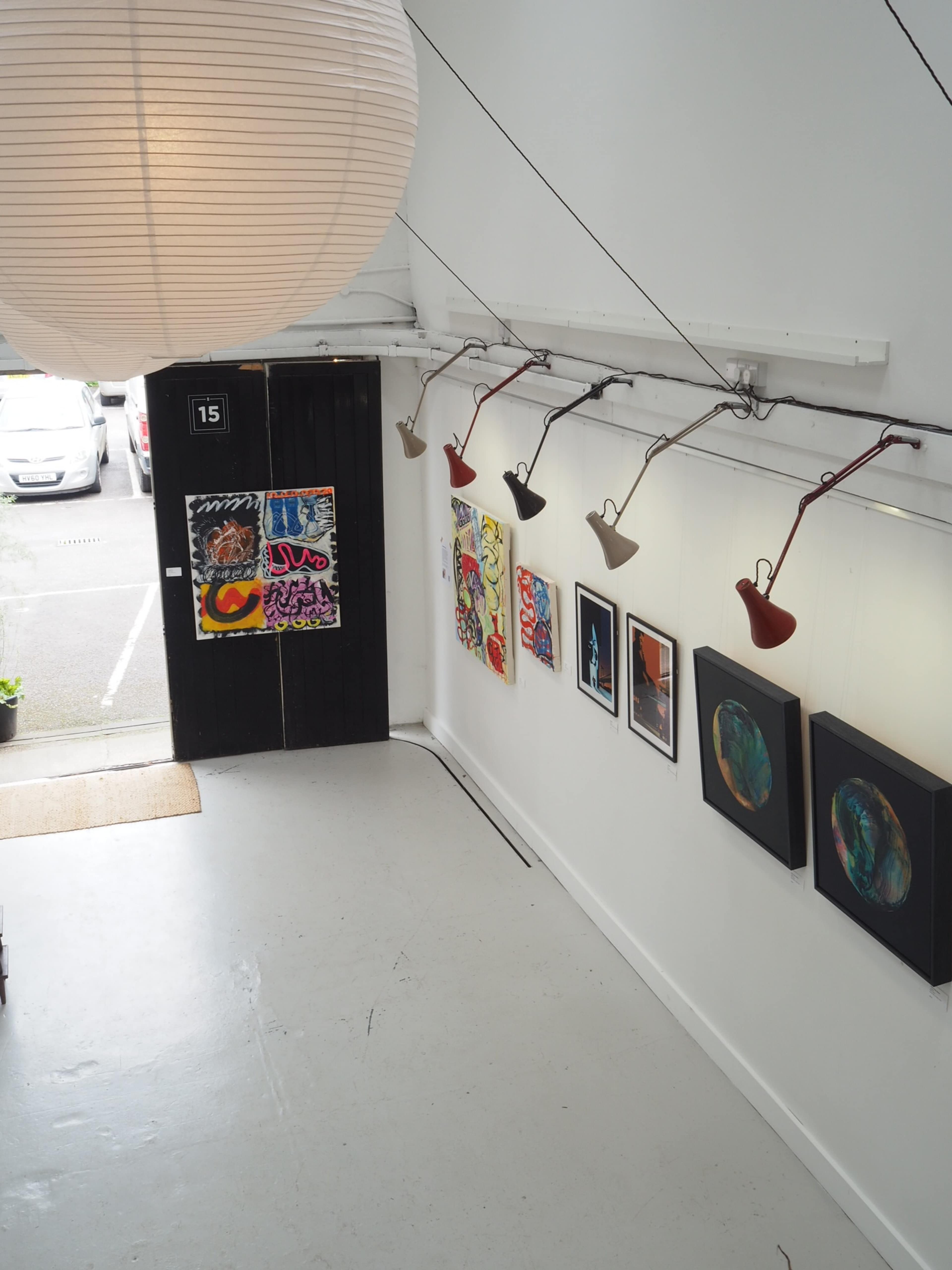 The image shows an art gallery with several framed artworks hanging on white walls, a large round lamp overhead, and a door leading outside.
