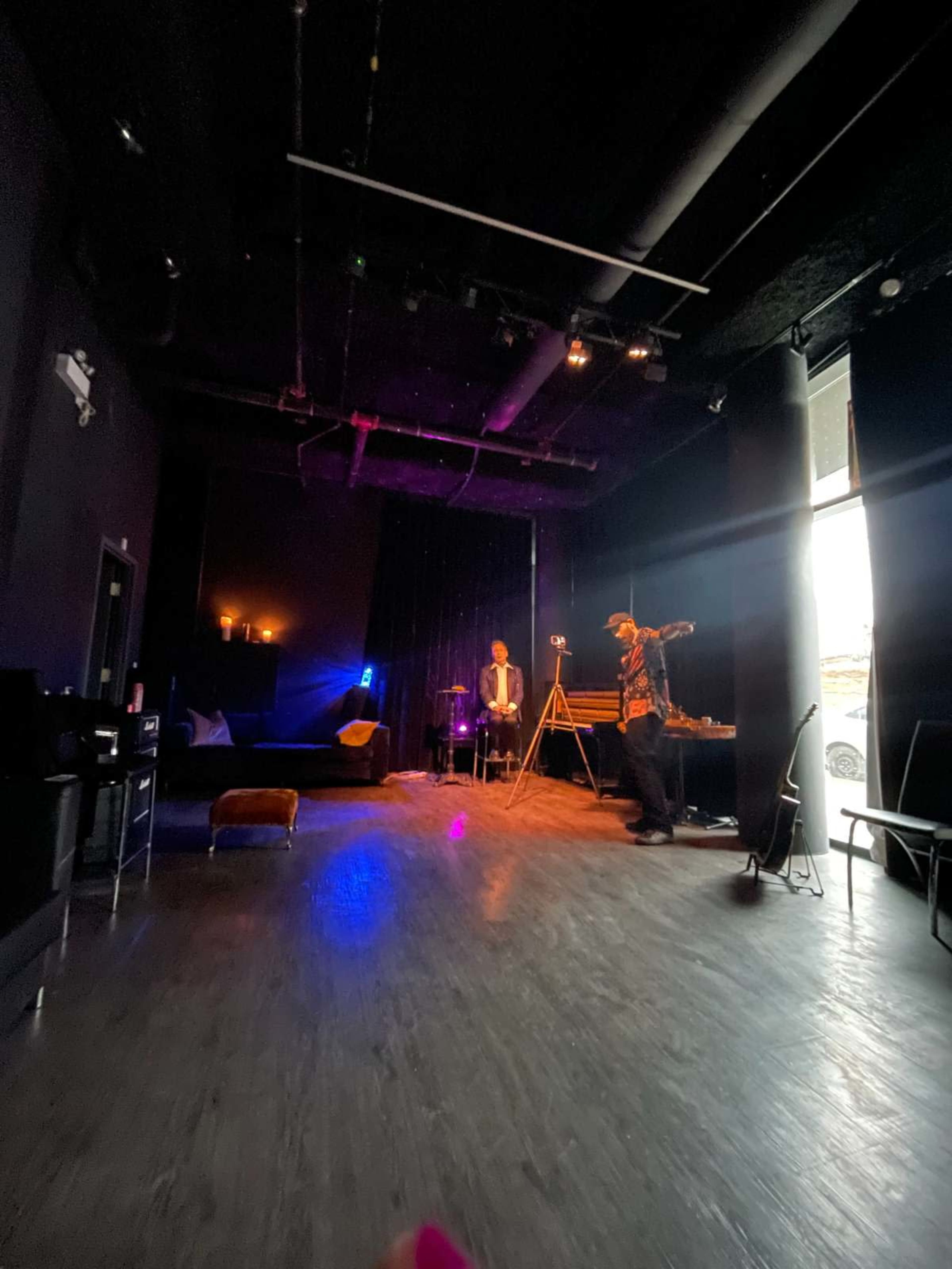 The image shows a dimly lit performance space with a performer on stage, a seated audience member, and a window letting in natural light.