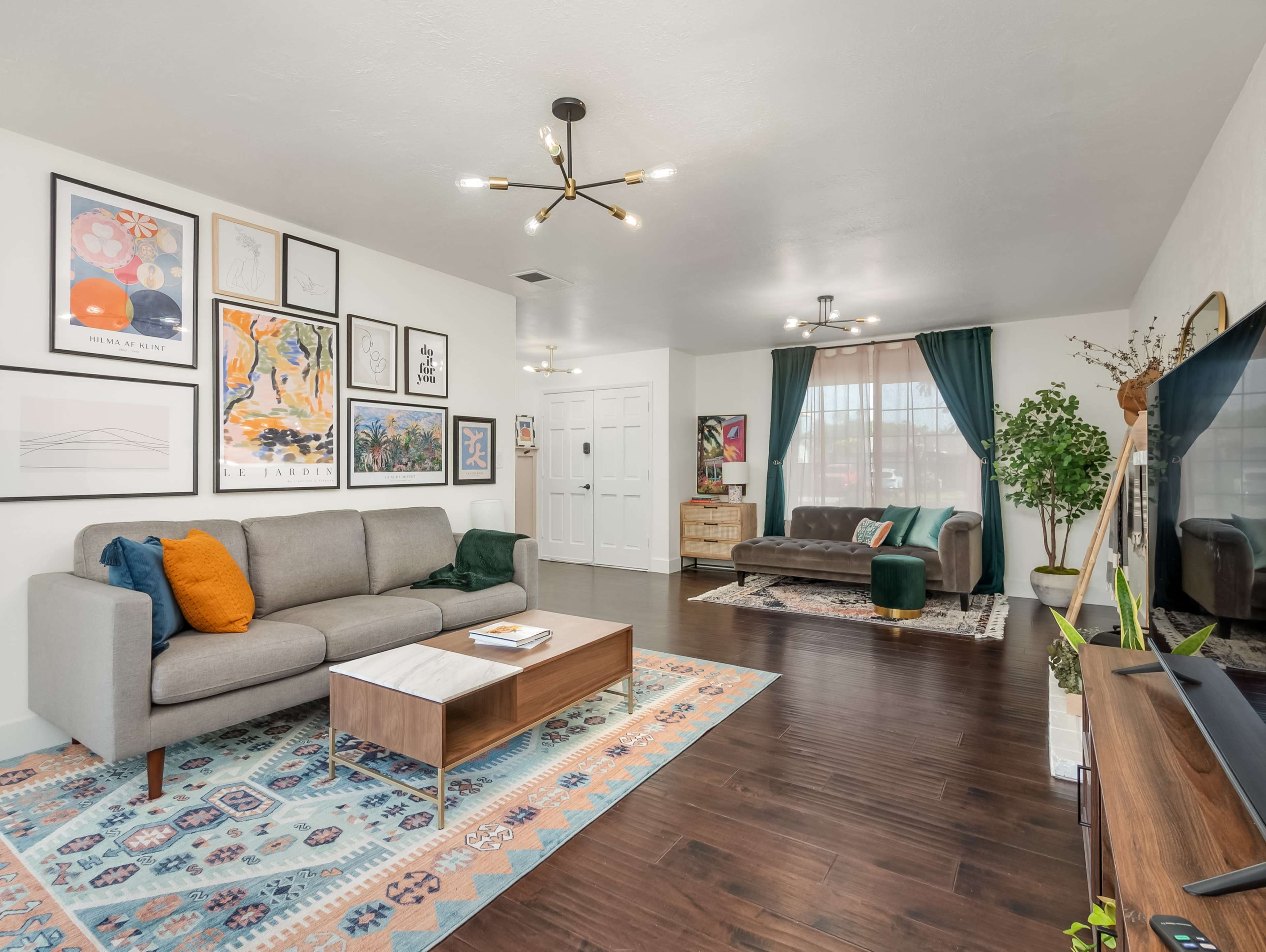 A modern living room features a gray sofa, a green chair, and various framed artworks on the walls, with a light-colored rug on dark wooden flooring.