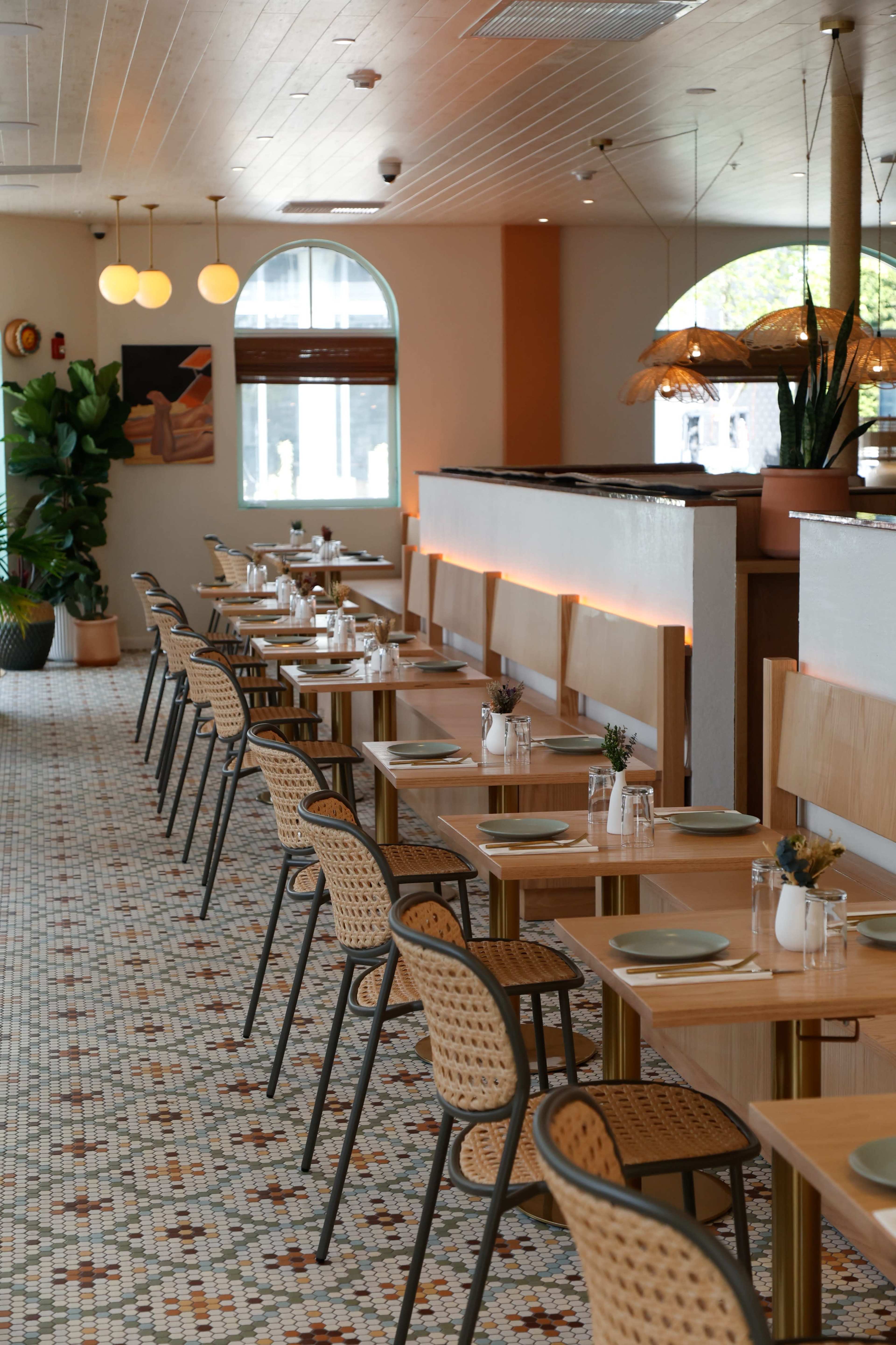 The image shows a modern restaurant interior with neatly arranged wooden tables and chairs, featuring a patterned tile floor and warm lighting.