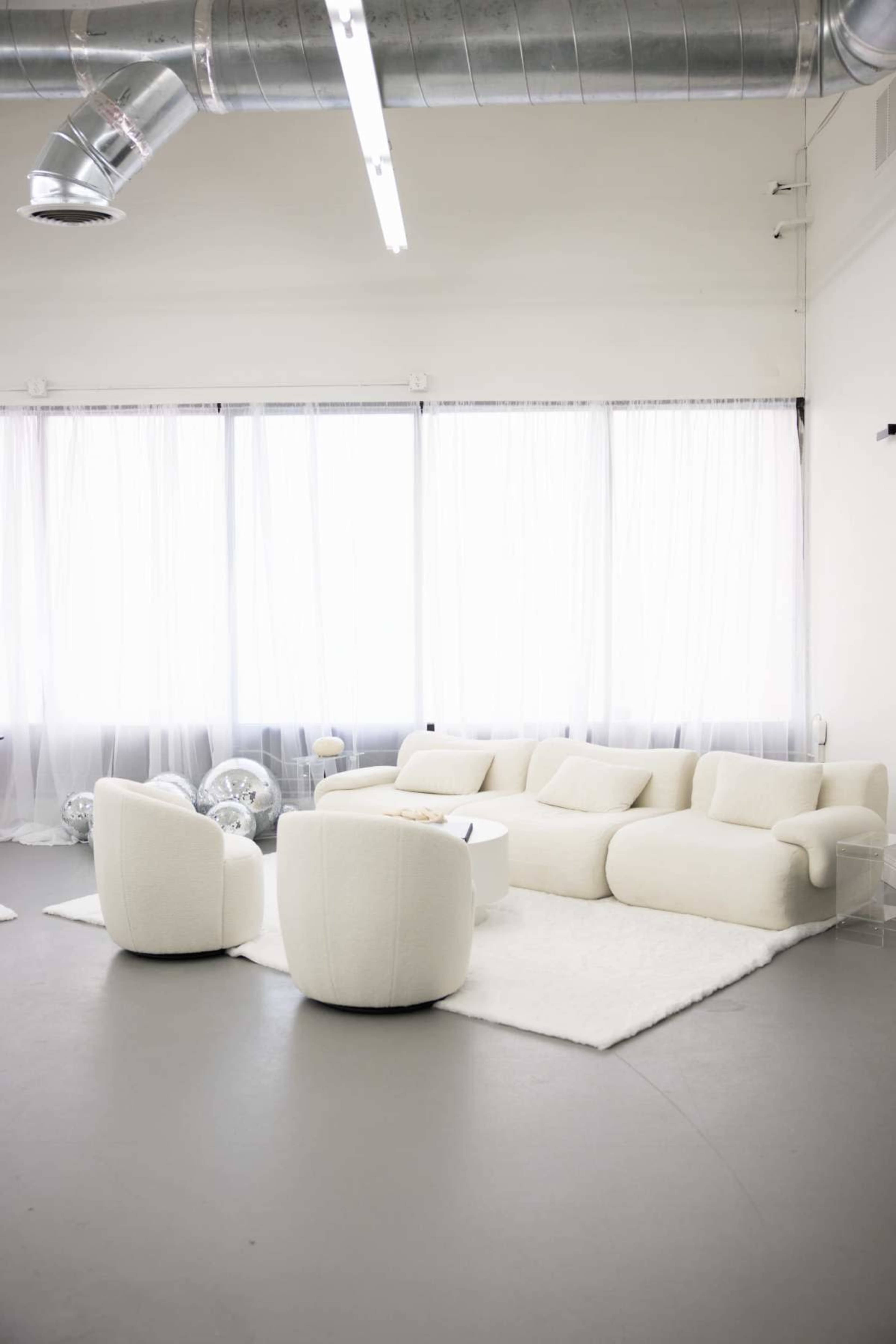 The image features a modern living room with a white sectional couch, two round upholstered chairs, and a coffee table on a soft rug, all situated under large windows with sheer curtains.