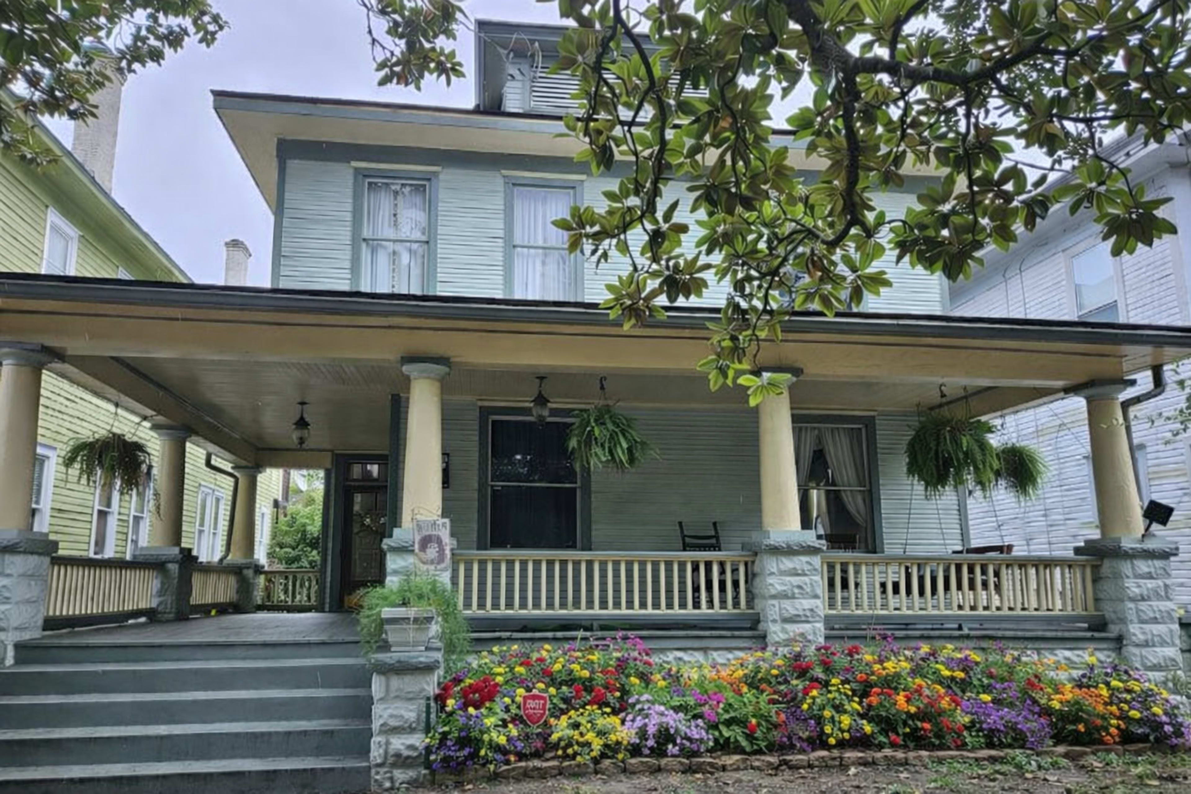A two-story blue wooden house with a large front porch adorned with hanging plants and vibrant flower beds.