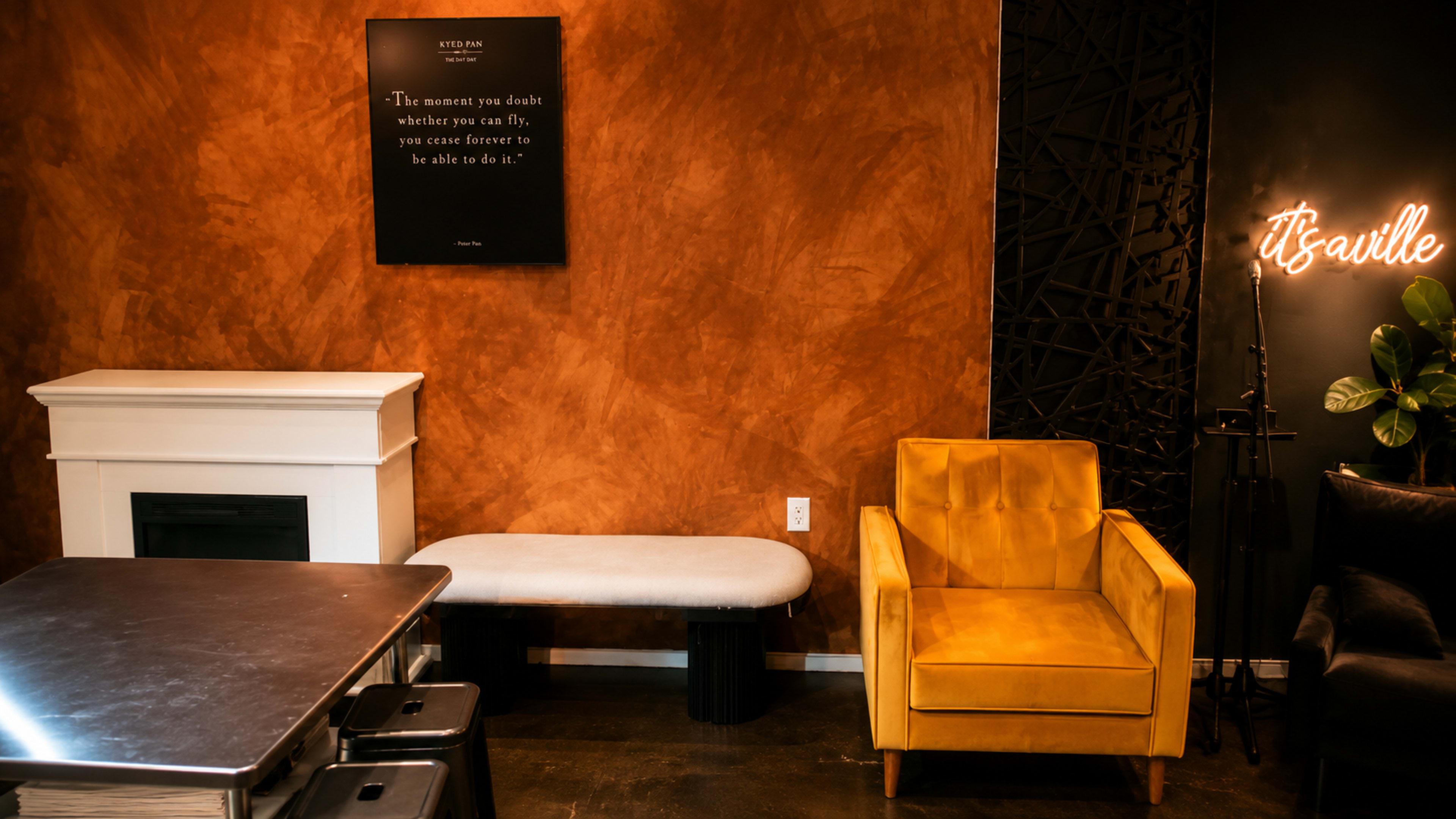 The image shows a modern interior featuring a yellow armchair, a white bench, a fireplace, and decorative wall art with a motivational quote.