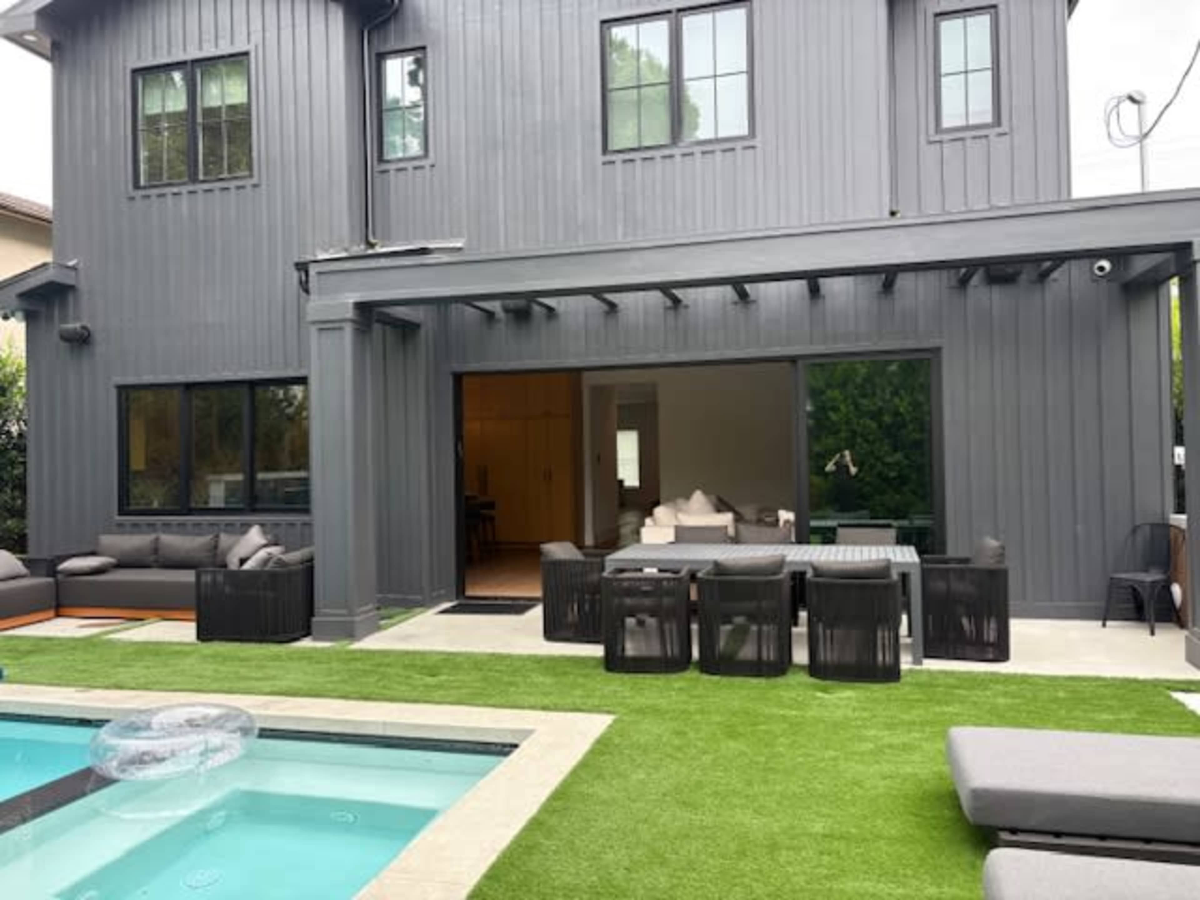A modern house with gray-clad exterior features a backyard patio that includes a pool, seating area, and dining set on artificial grass.