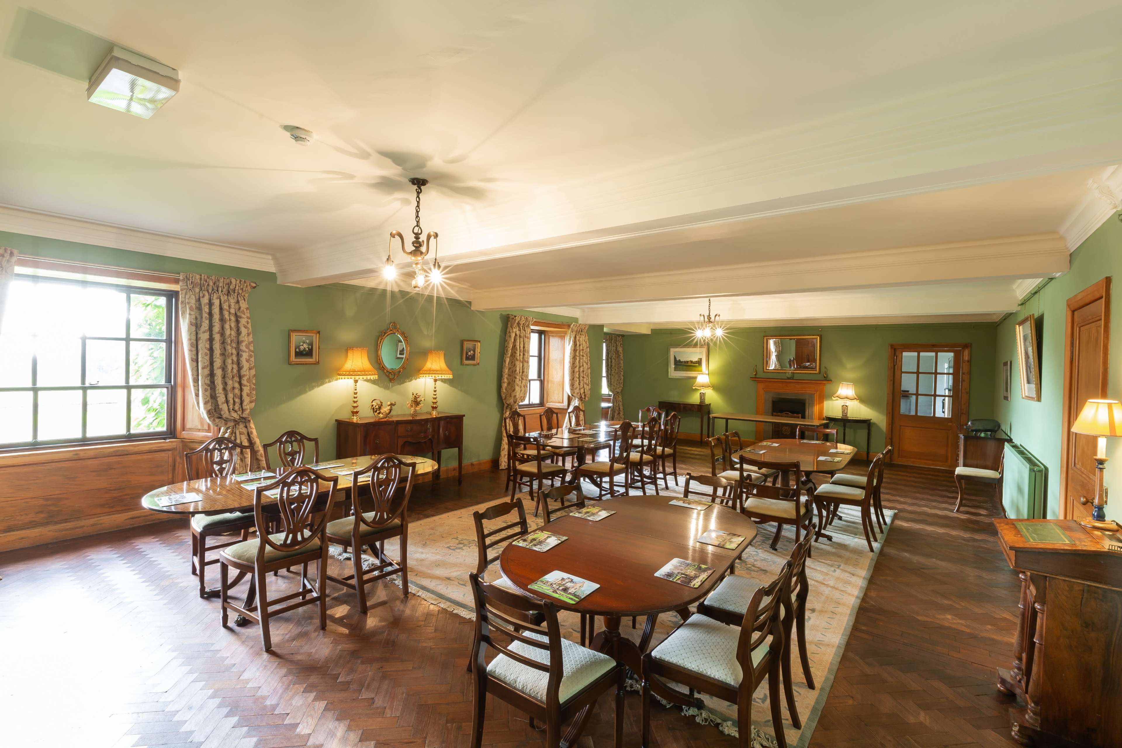The image shows a spacious dining room with wooden furniture, decorative artwork on the walls, and large windows allowing natural light to enter.