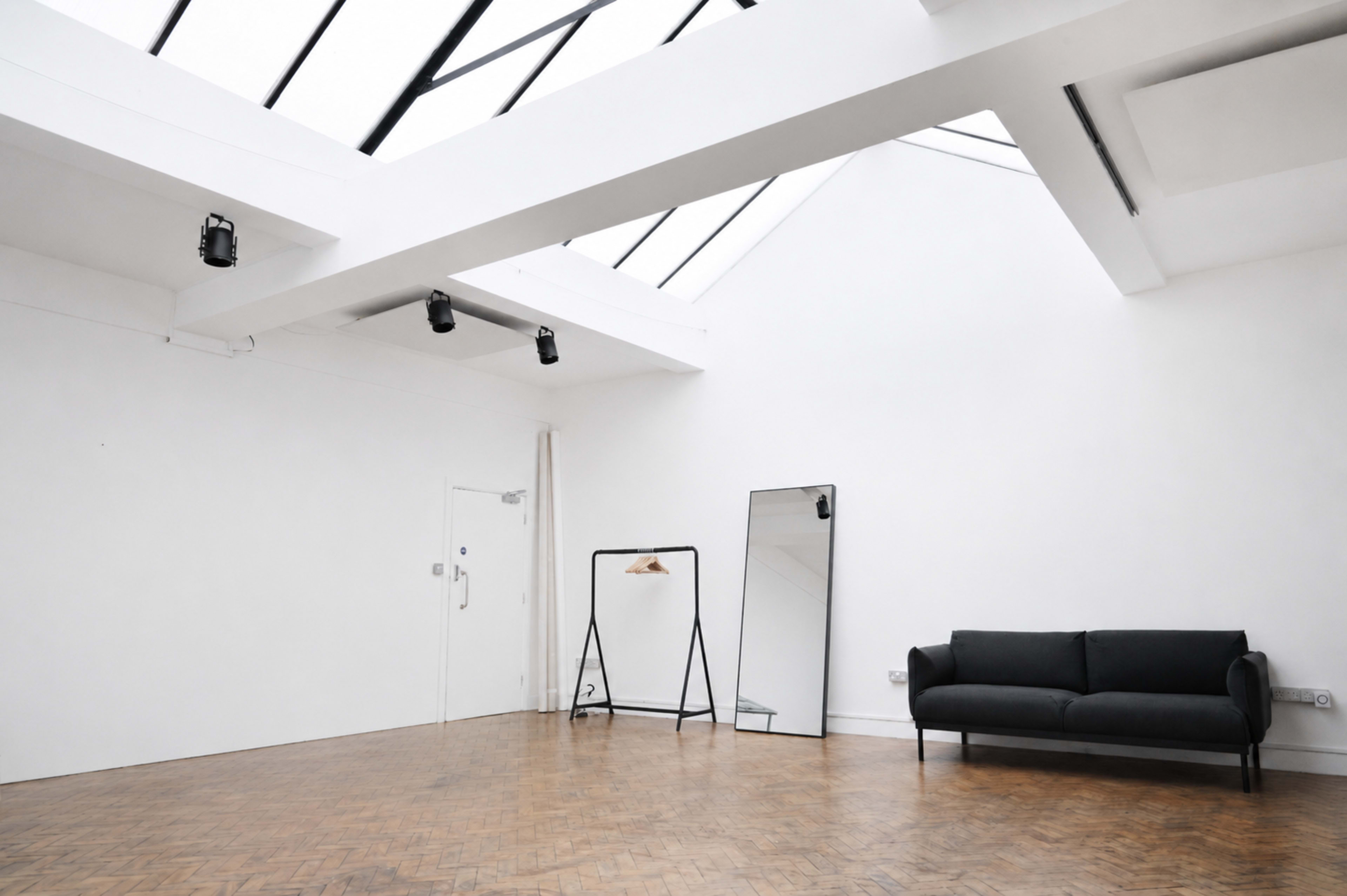 The image shows a minimalist interior space with a high ceiling, large windows, a black couch, and a mirror on a stand.
