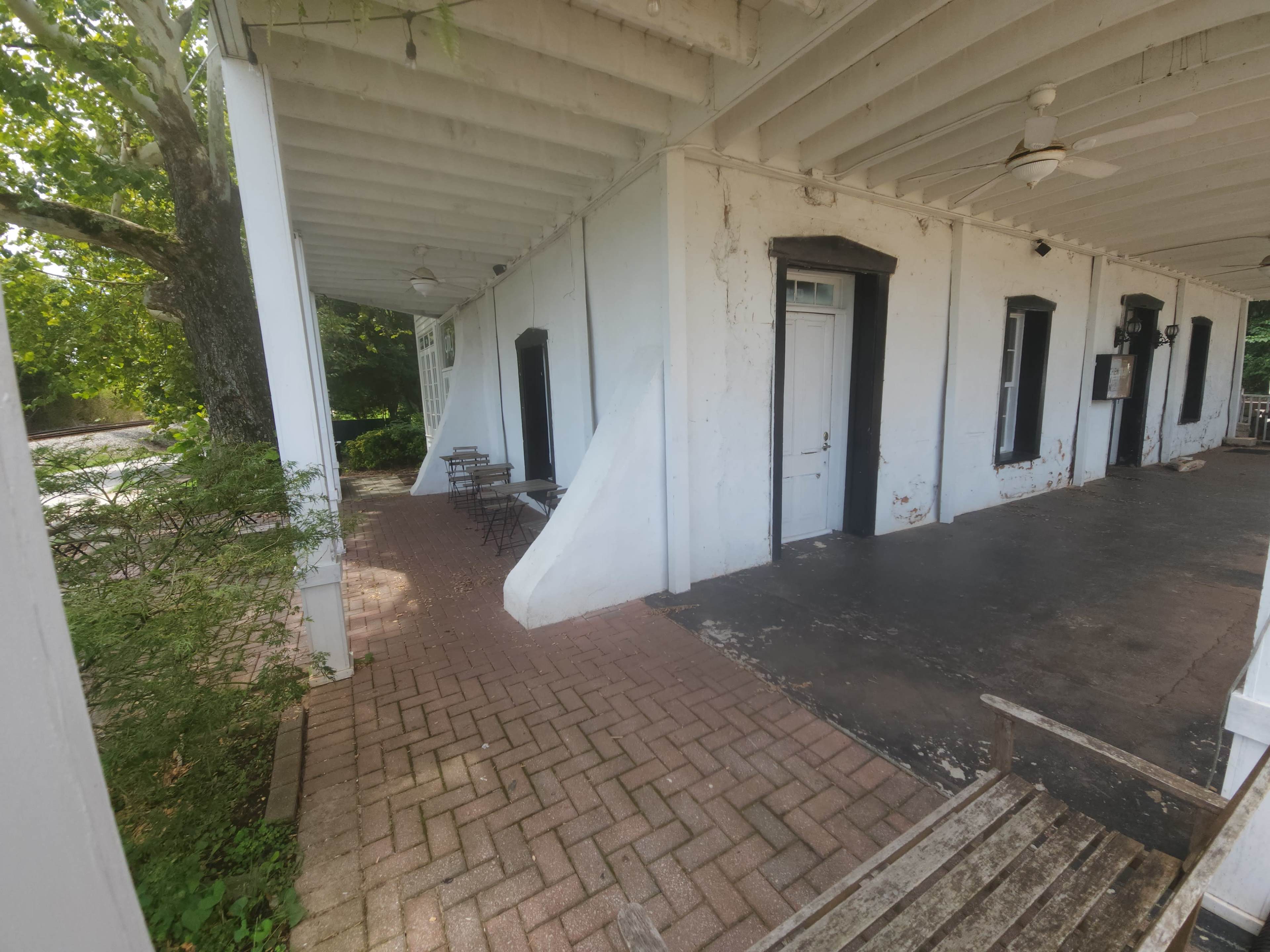 The image shows a covered porch area with white walls, a brick pathway, and a few small tables and chairs under the shade of trees.