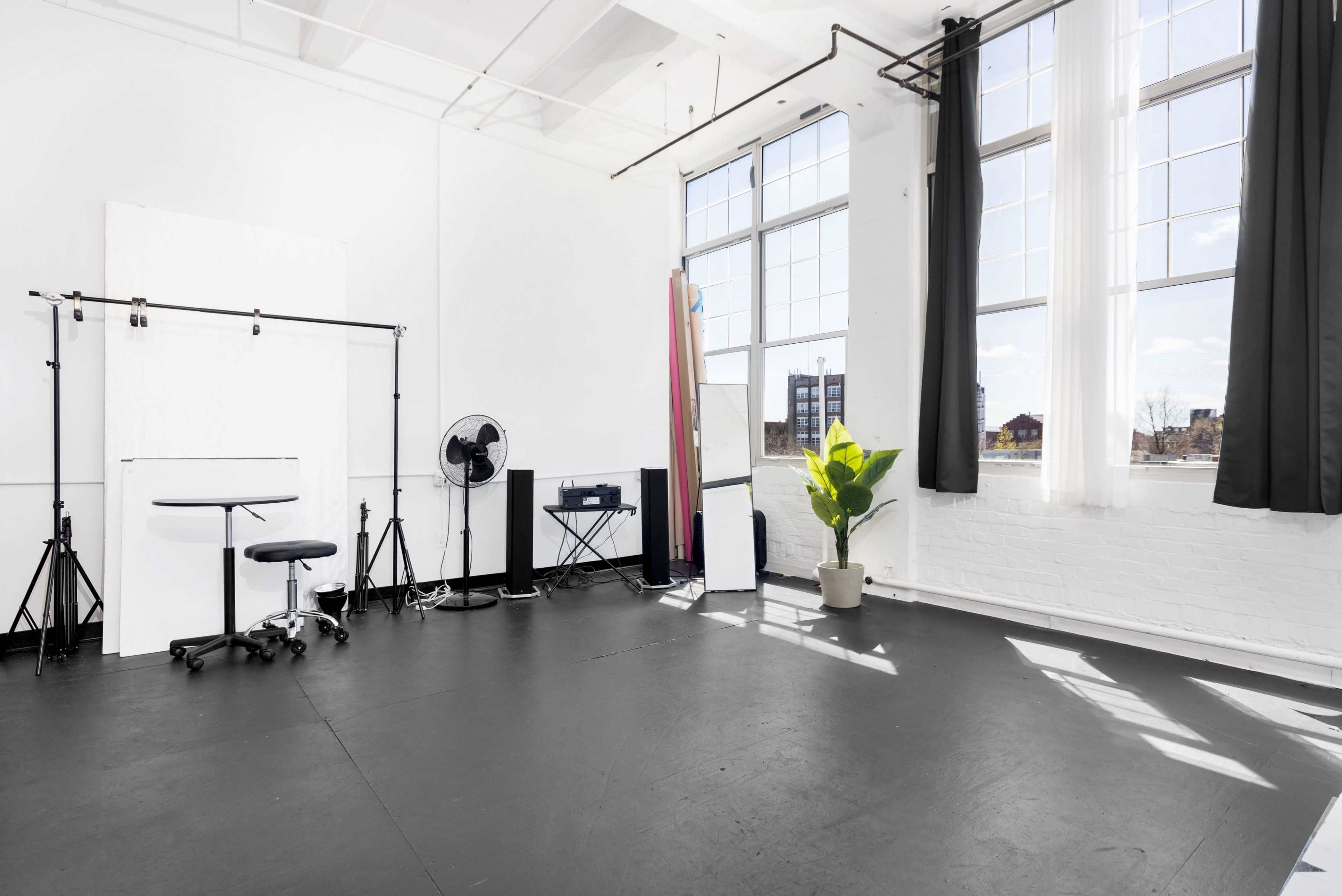 A bright, empty studio space with large windows, a fan, a mirror, a small table, and a potted plant.