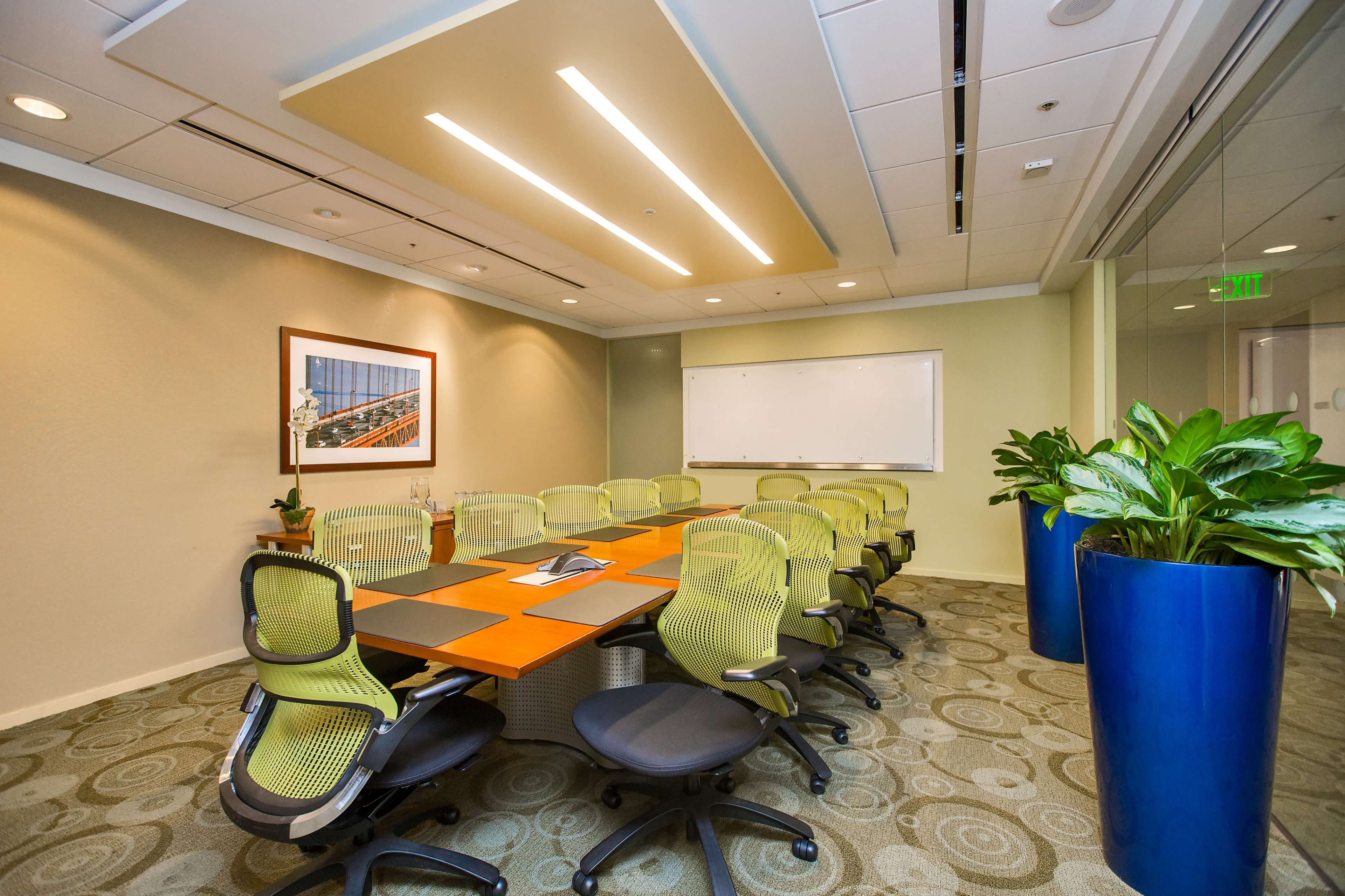 A modern conference room with a long table surrounded by ergonomic chairs, a whiteboard on one wall, and large potted plants in the corners.
