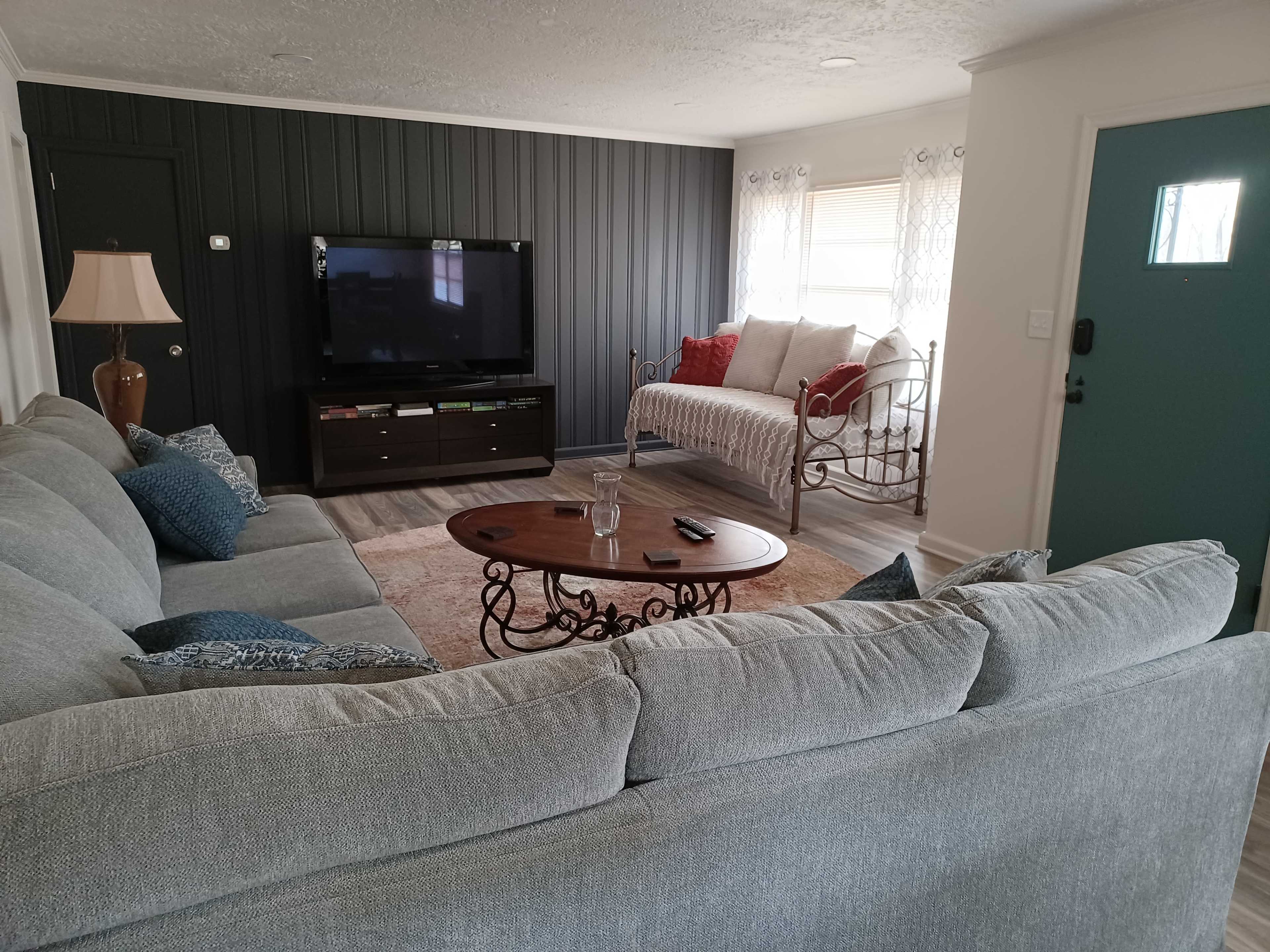 A living room features a gray sectional sofa, a coffee table, a television on a dark cabinet, and a daybed near a teal-colored door.