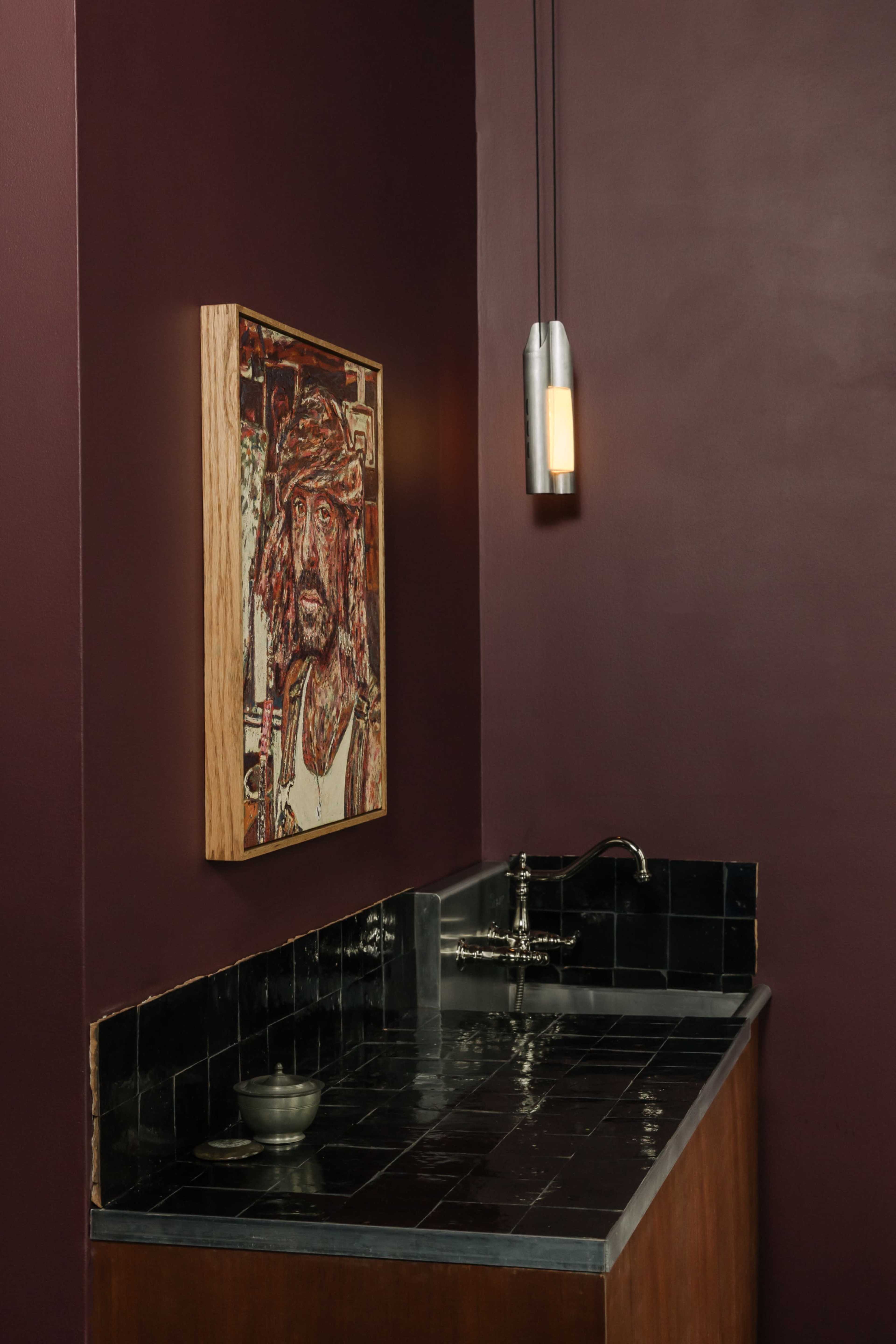 A dark-hued bathroom features a wooden-framed portrait on the wall above a sleek black countertop with a sink and a small bowl.