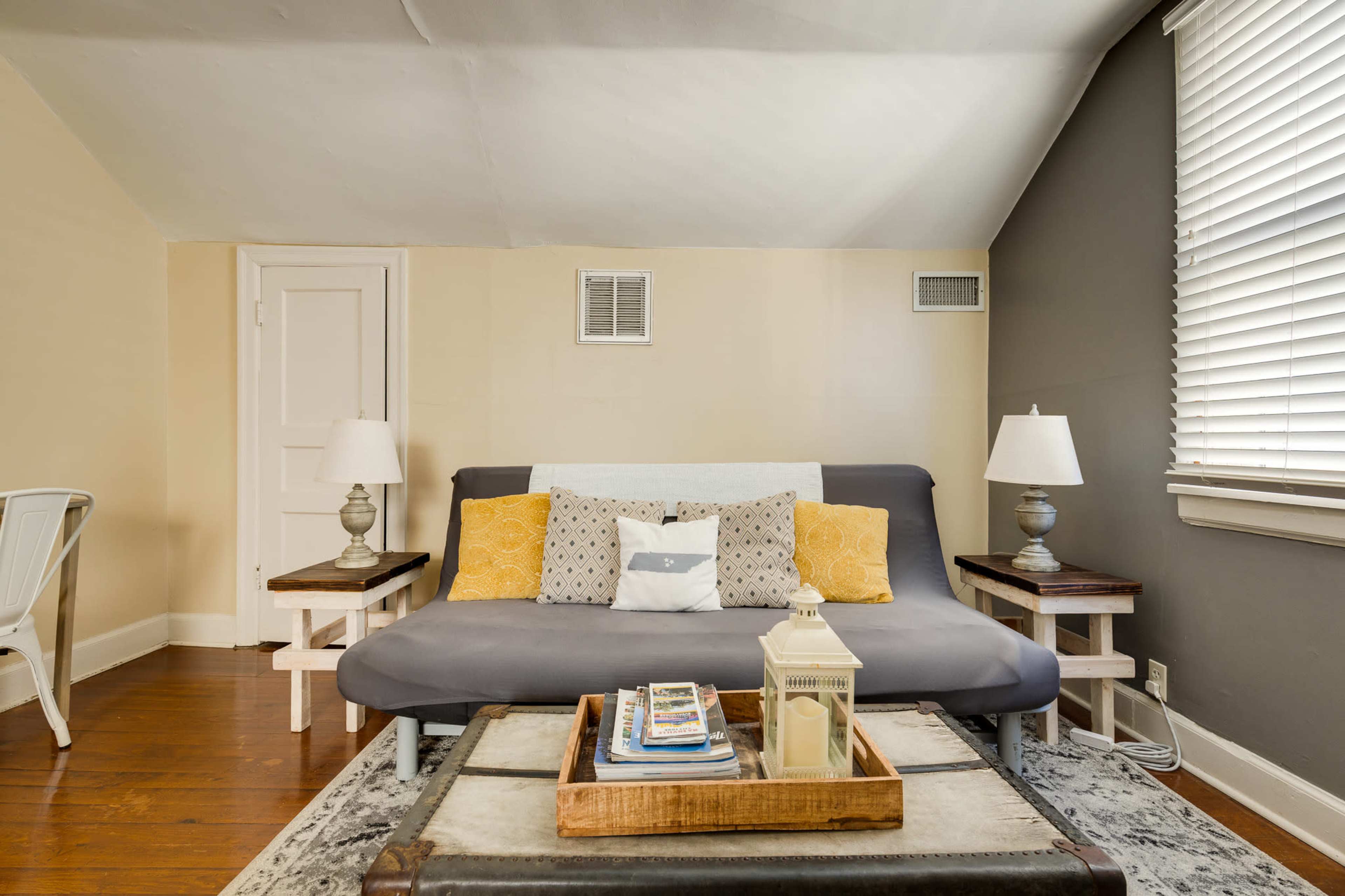 The image shows a cozy living space featuring a gray sofa, decorative pillows, two lamps on side tables, a coffee table with books, and wooden flooring.