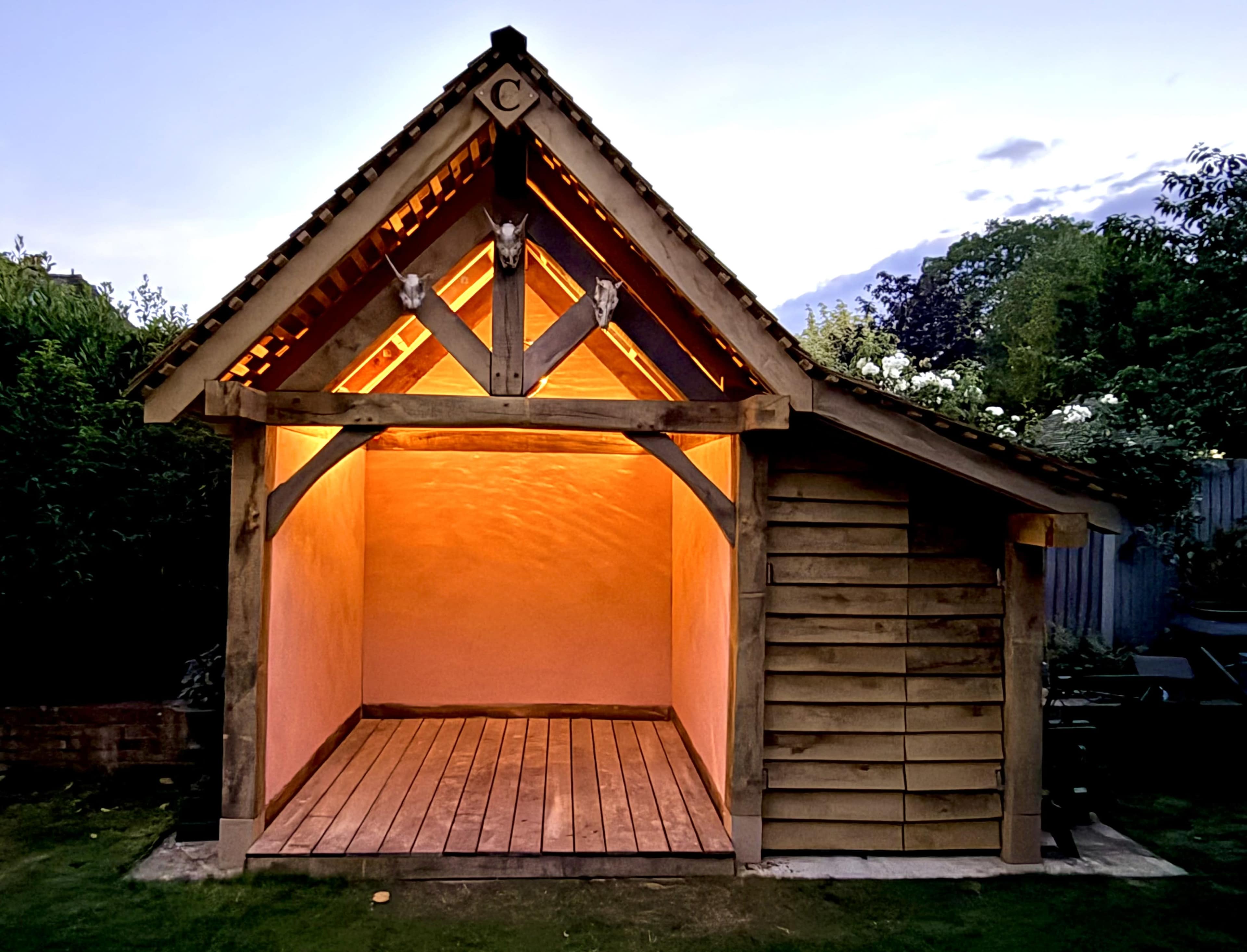 A wooden garden shed with a peaked roof is illuminated from within, showcasing its empty interior against a twilight sky.