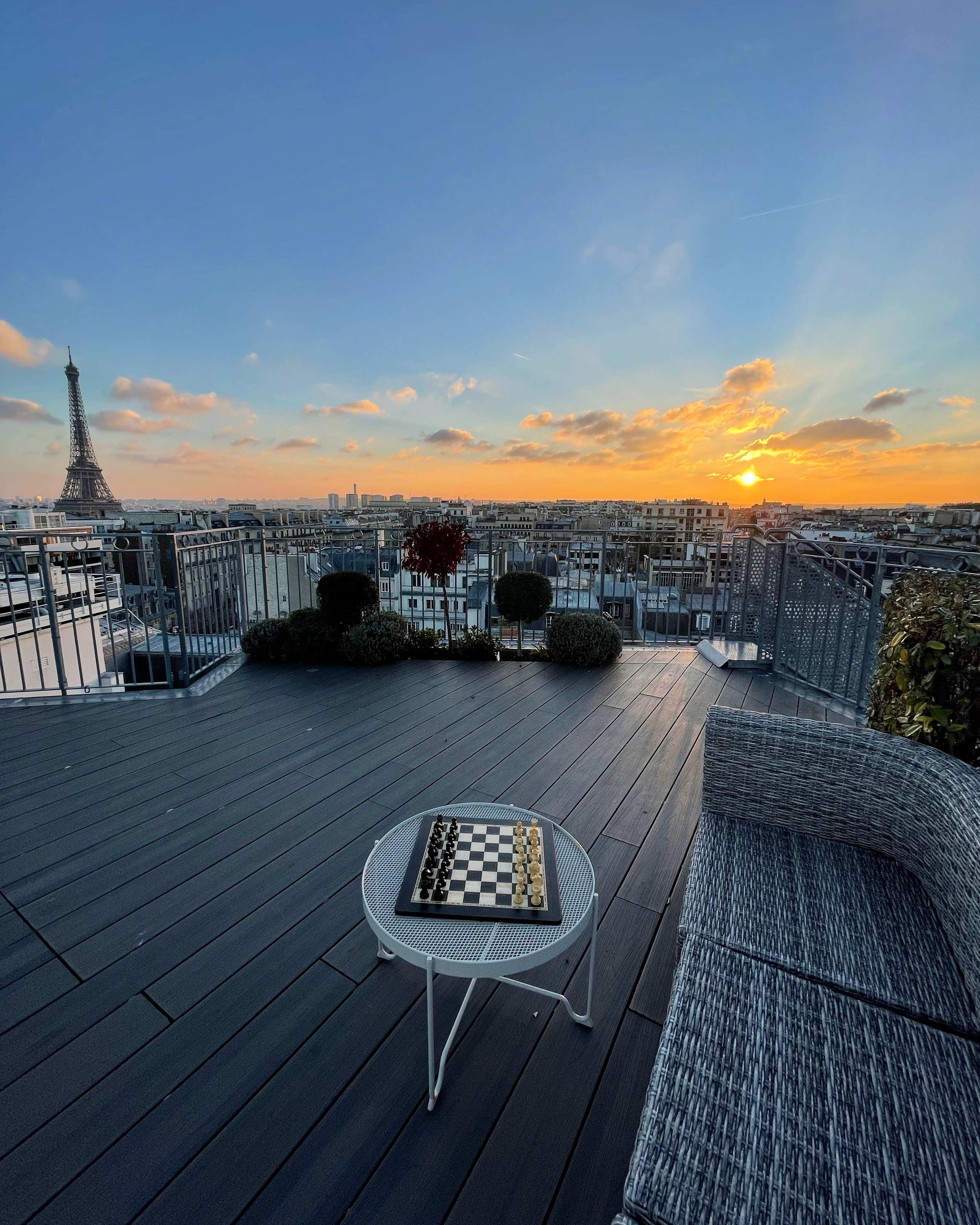 A chessboard sits on a table on a rooftop terrace overlooking the Paris skyline at sunset, with the Eiffel Tower visible in the background.