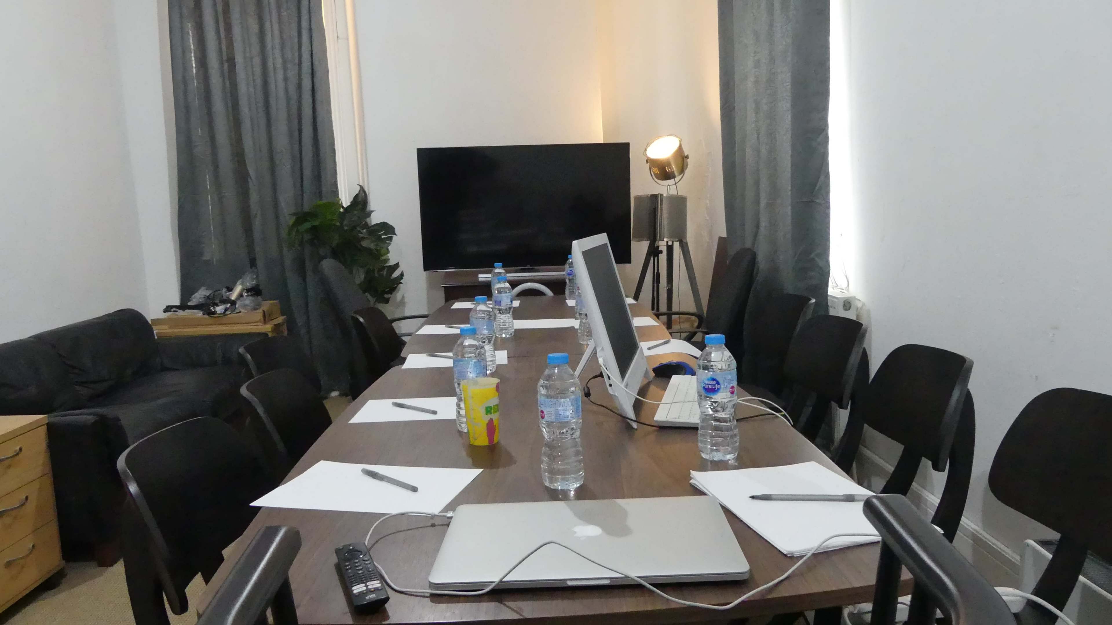 A conference room is set up with a long table, several chairs, bottles of water, notepads, and electronic devices, including a computer and a television.