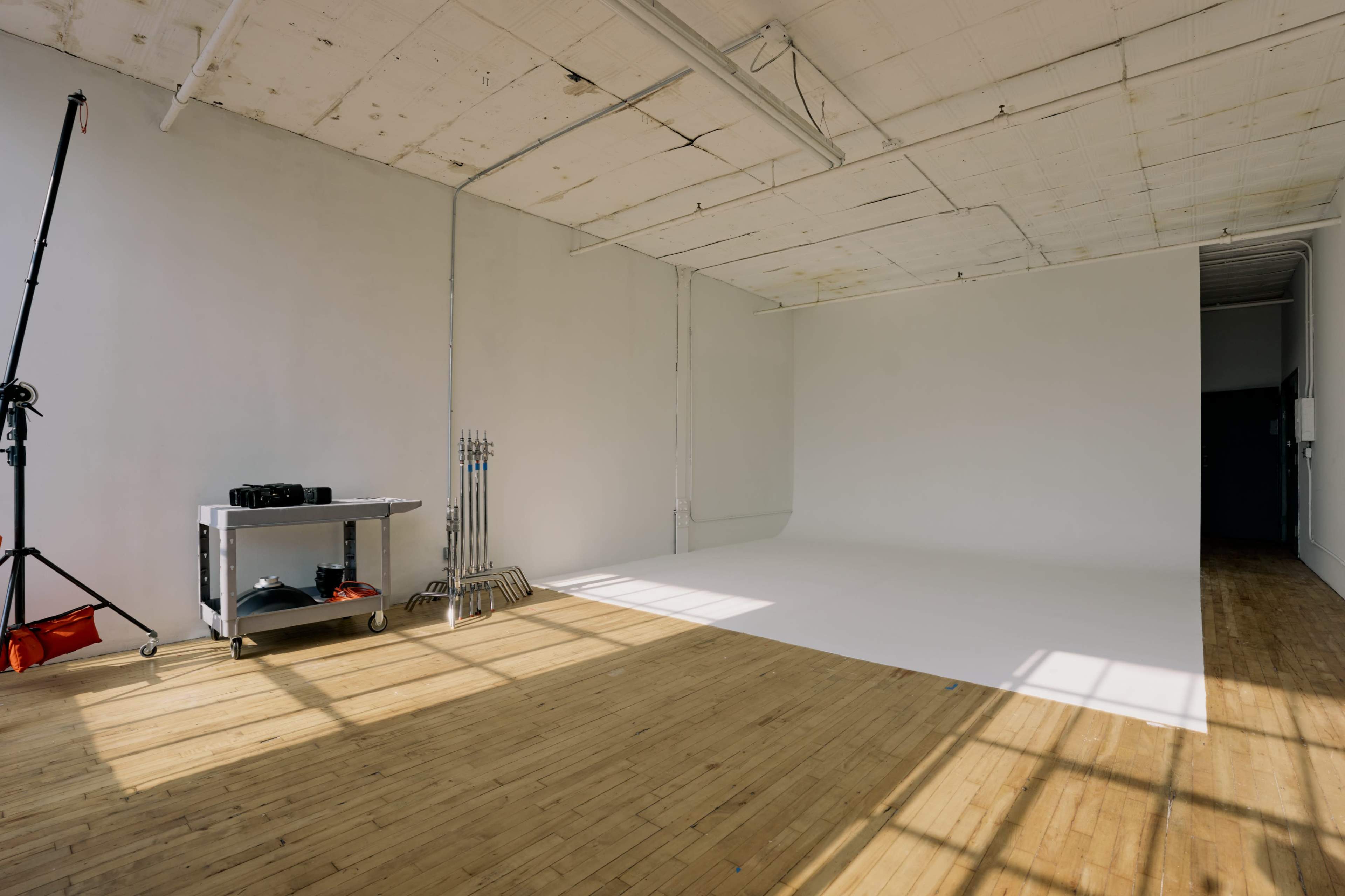 The image shows a spacious, empty studio with a blank white wall, wooden flooring, and a few photography equipment items on a cart.