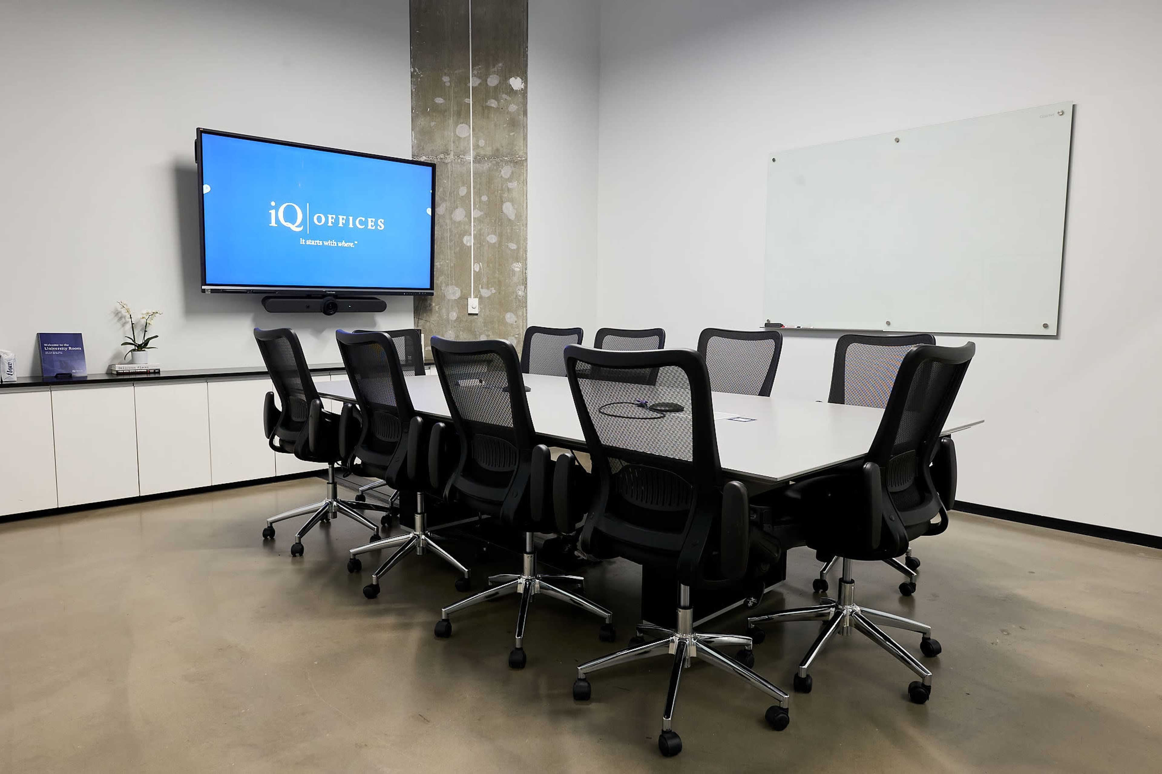 The image shows a modern conference room with a large table surrounded by ergonomic chairs, a wall-mounted screen displaying a logo, and a whiteboard for presentations.
